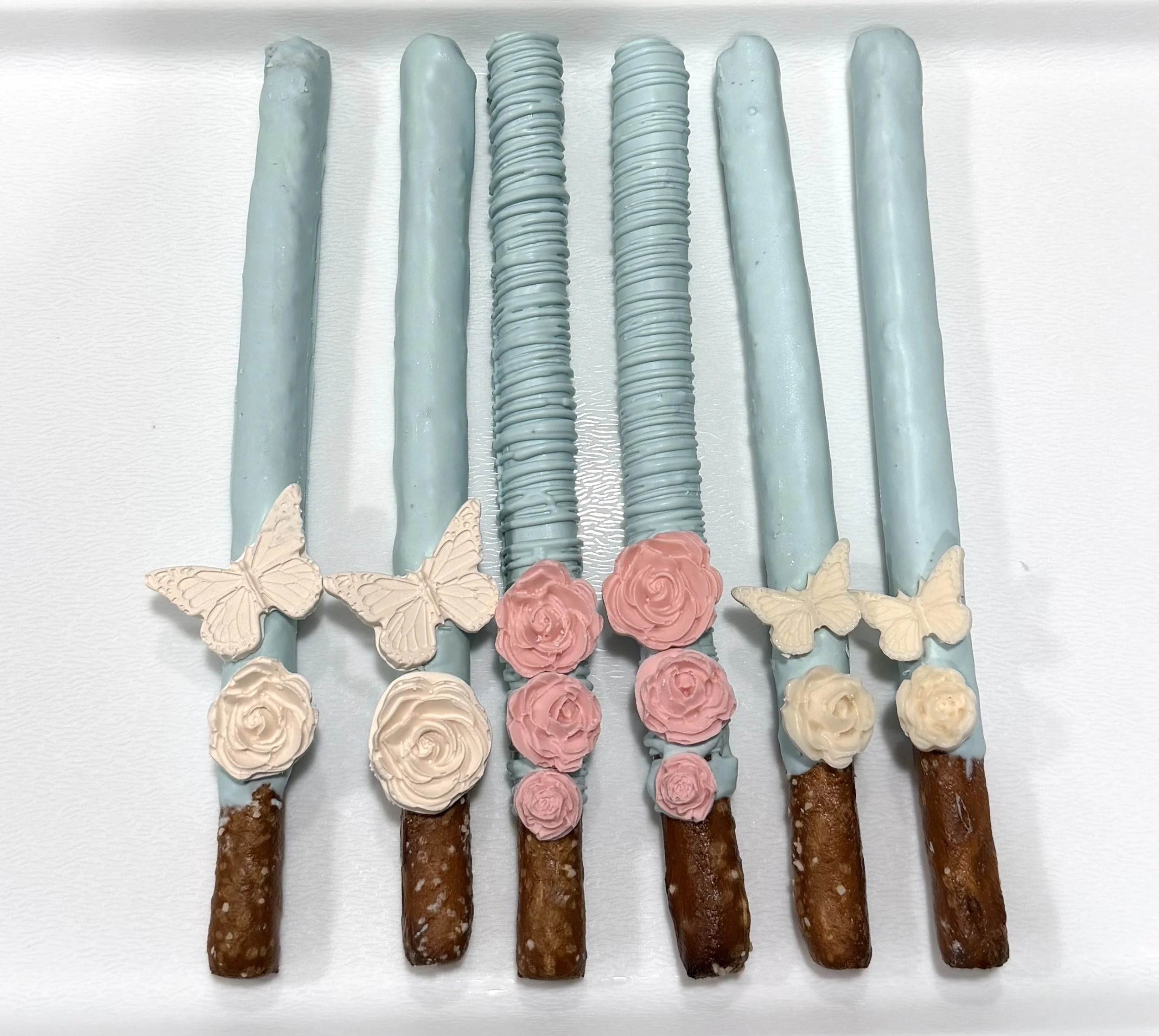 A row of six ice cream cones with pink, cream, and blue icing, decorated with butterfly and rose-shaped toppers, placed on a white surface.