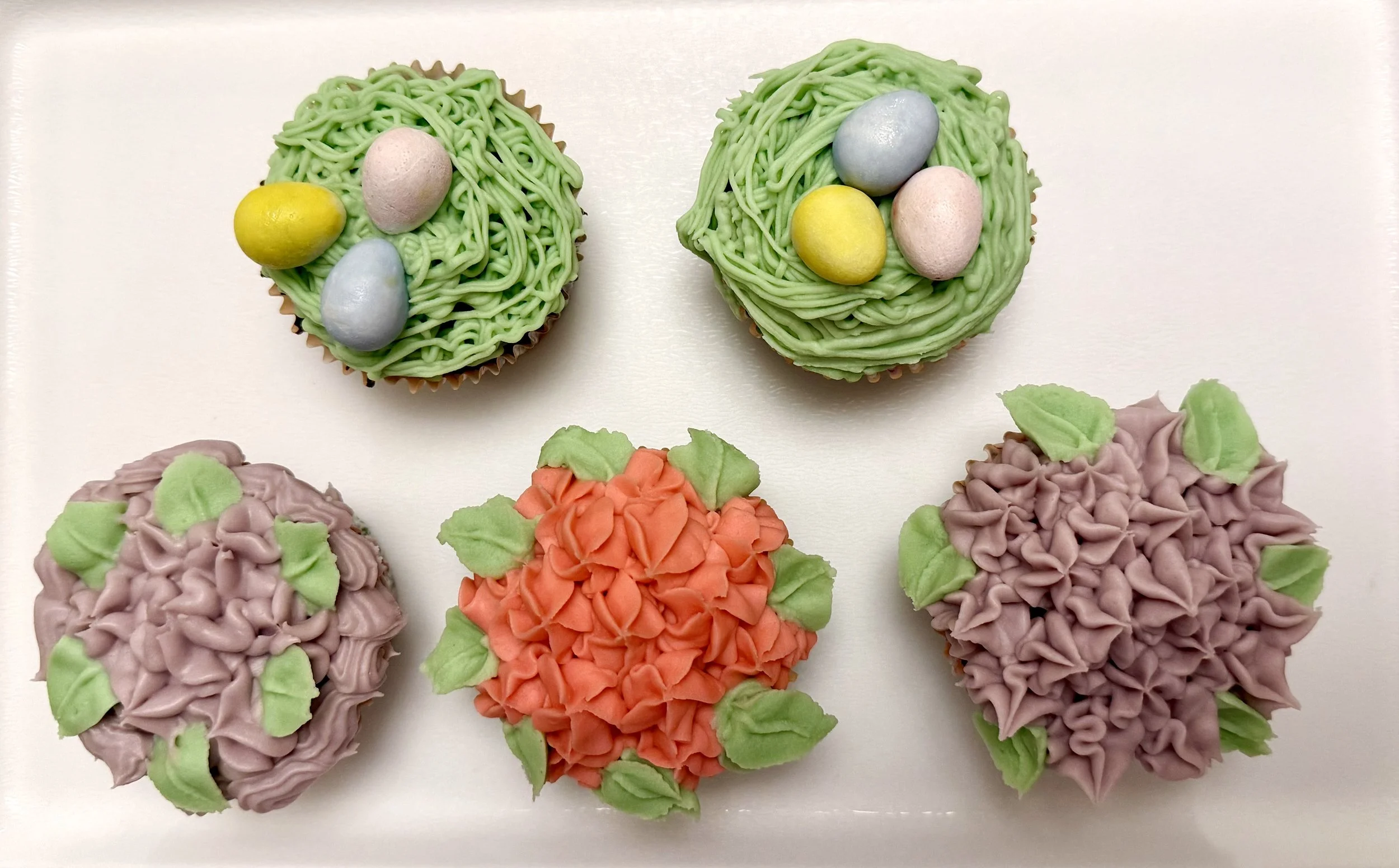 Decorative cupcakes with pastel-colored frosting designed to look like Easter eggs and flowers, arranged on a white surface.