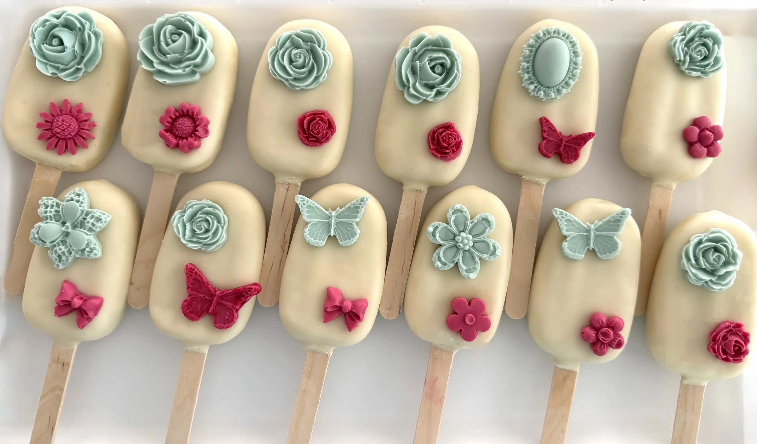 A row of white ice cream bars decorated with colorful edible flowers, butterflies, and bows on wooden sticks.