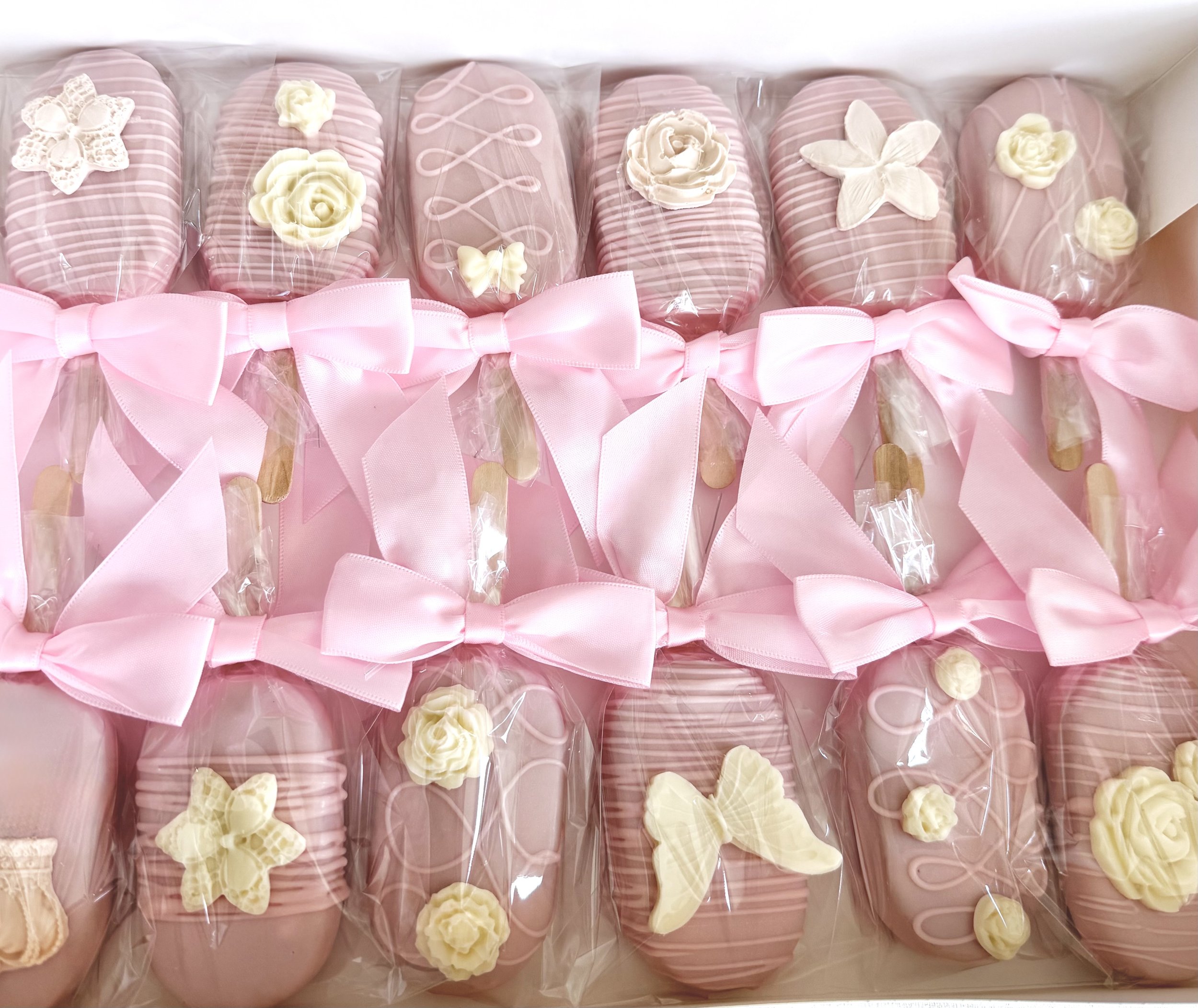 A collection of pink and white decorated cake pops with bows and floral decorations, wrapped in clear plastic.