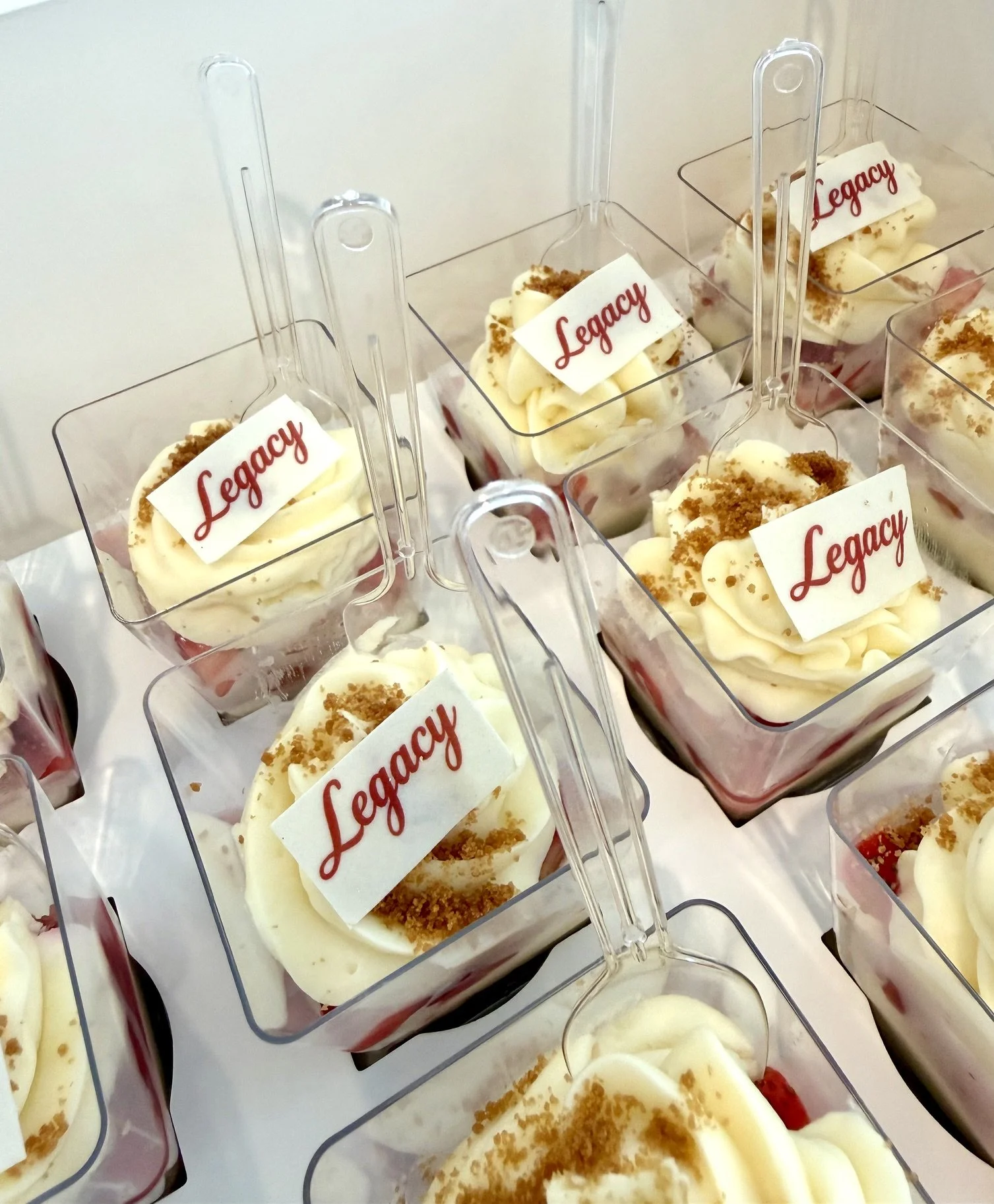 Strawberry shooters with cream cheese, crumble Biscoff topping, and 'Legacy' written on a white chocolate piece, arranged in clear containers.