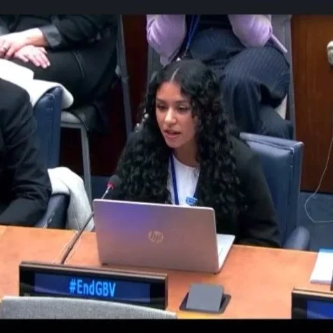 Young woman (Neisha Hussain) with long curly hair speaking into a microphone at a formal meeting or conference, sitting at a table with a laptop in front of her, with others seated around her.
