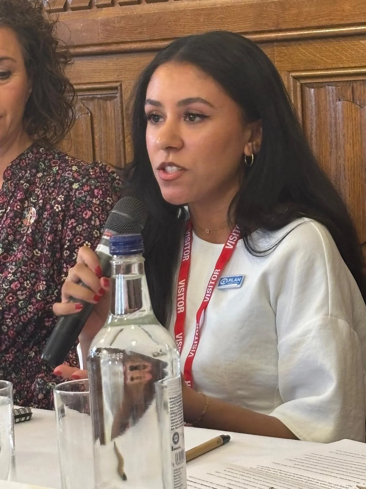 Young woman (Neisha Hussain) with black hair, wearing a white shirt, holding a microphone, and sitting at a table with documents and a water bottle, speaking during an indoor event.