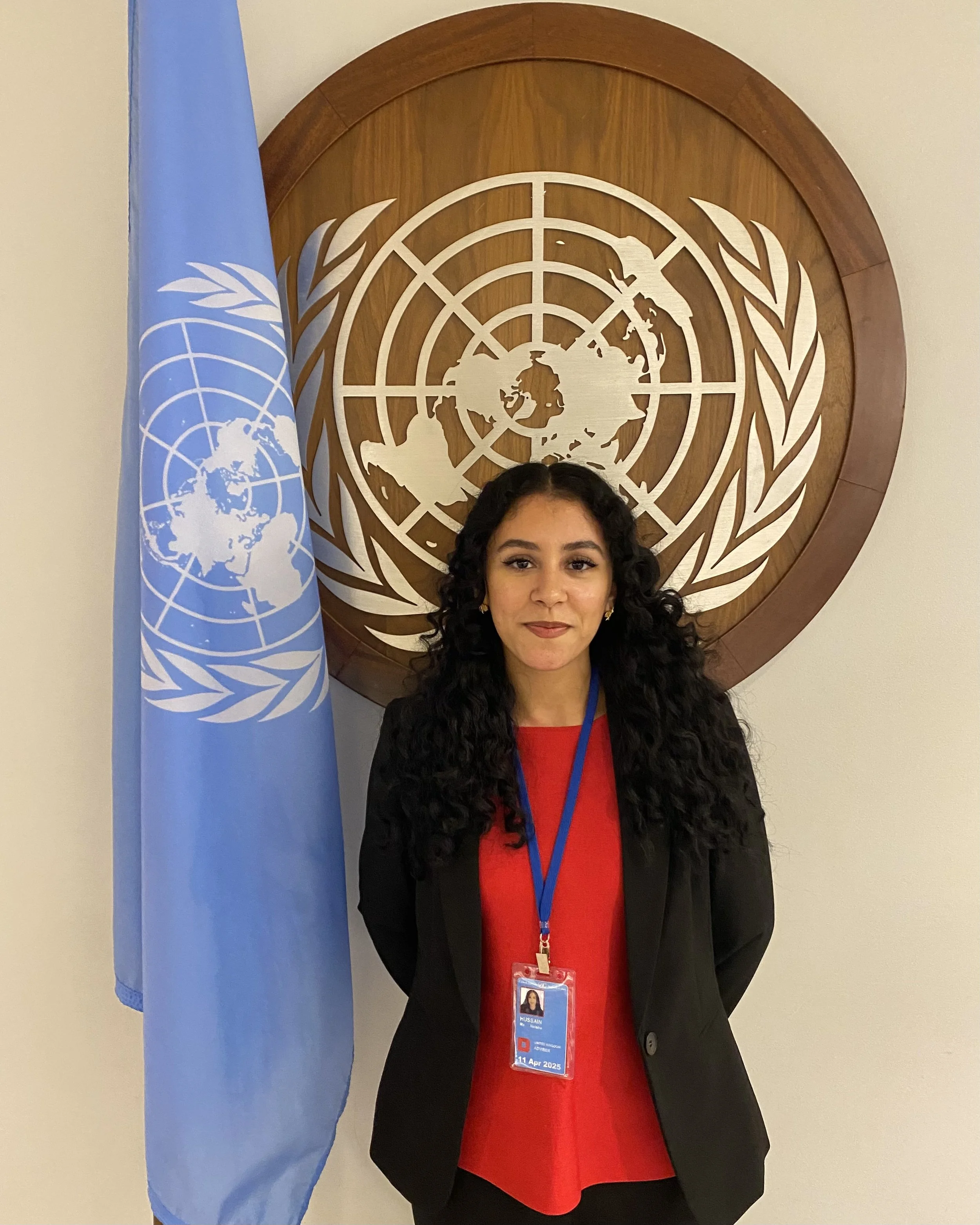 A woman (Neisha Hussain) with long curly black hair wearing a black blazer and a red top, standing in front of the United Nations emblem and flag.