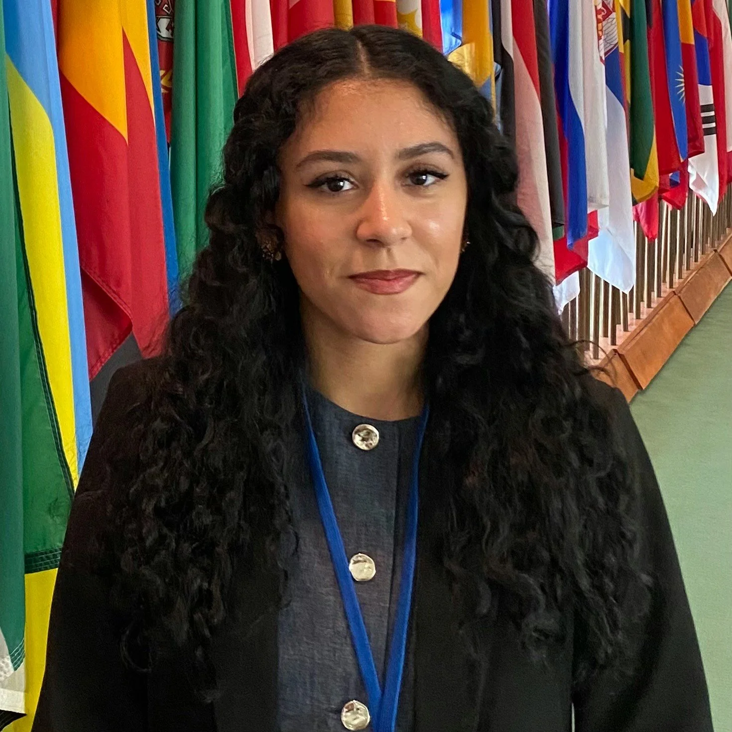 A woman (Neisha Hussain) with curly black hair wearing a dark jacket and a blue lanyard with a badge, standing in front of a row of international flags.