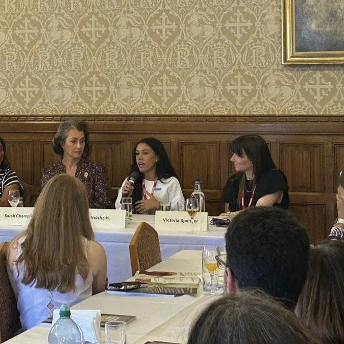 A panel of four women (including Neisha Hussain)seated at a table, with one woman speaking into a microphone. The setting appears to be a formal discussion or conference in a room with ornate wallpaper and wood paneling.