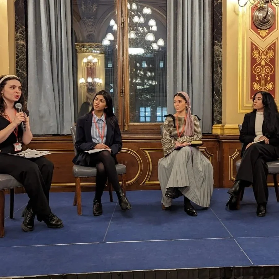Four women sitting on chairs on a stage in front of a large mirror, participating in a panel discussion or interview in a formal set-up, with chandeliers and ornate decor in the background.