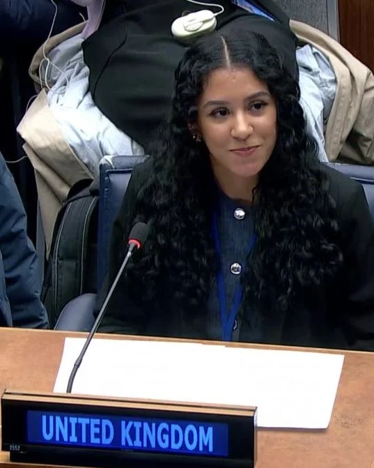 Young woman (Neisha Hussain) with long, curly black hair seated at a conference table, with a nameplate reading 'United Kingdom' in front of her, in a formal setting.