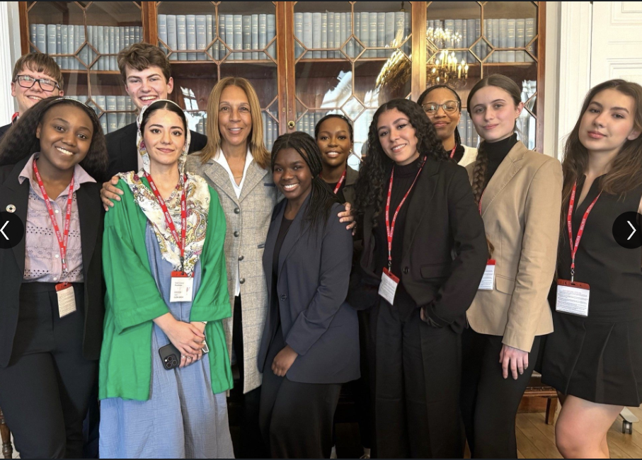 Group of ten diverse young women and one older woman posing together indoors, smiling, wearing business attire and conference badges, in front of a wooden bookshelf with glass doors filled with books.