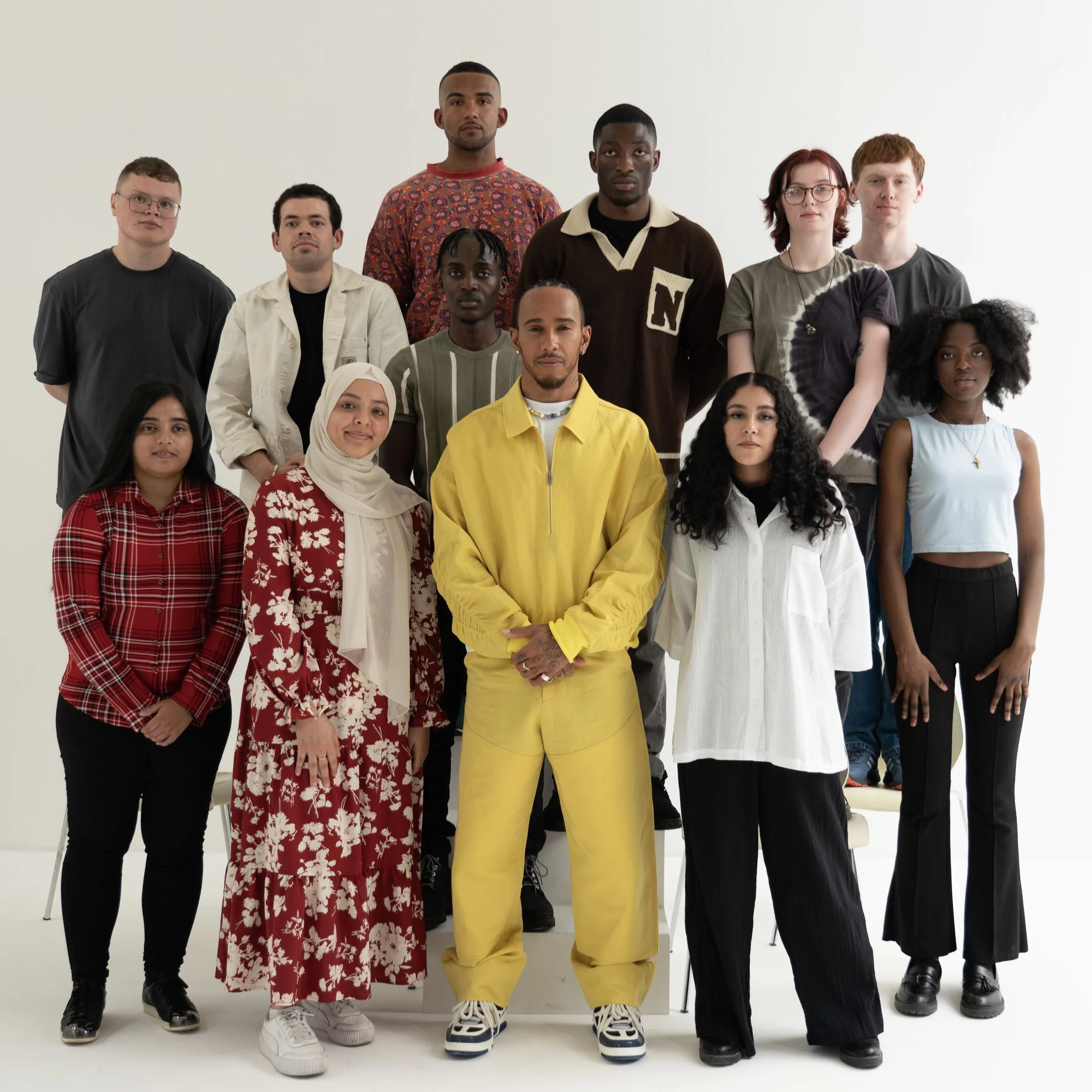 A diverse group of fifteen young people arranged in three rows poses against a white background with Lewis Hamilton