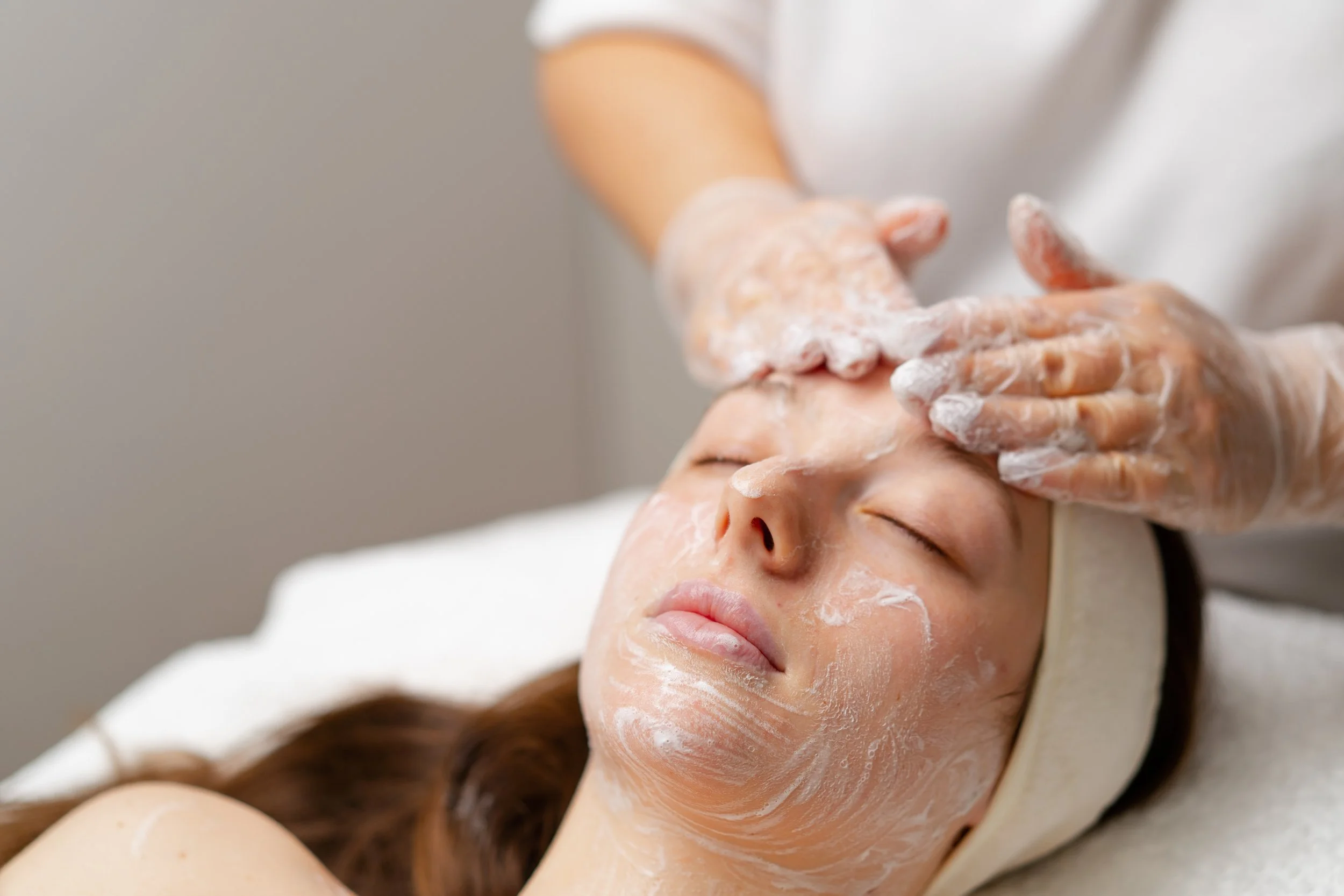 A woman receiving a facial treatment, lying down with her eyes closed, while a professional applies a white facial mask to her face.