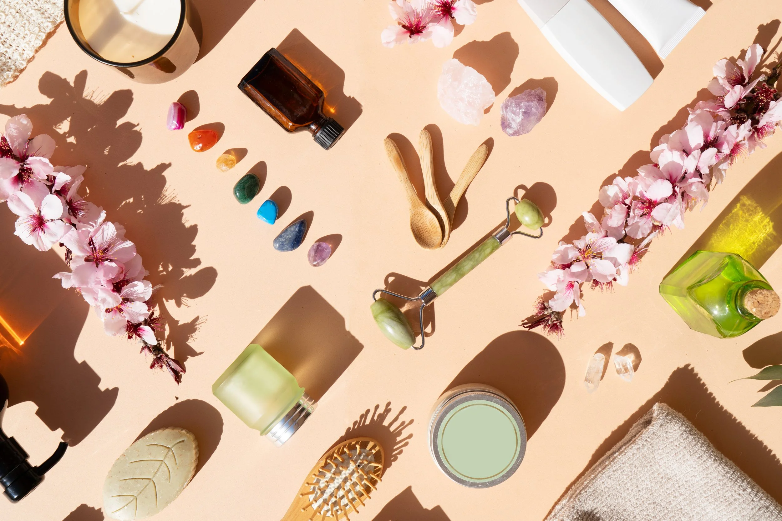 Flat lay of skincare and wellness items including a candle, small essential oil bottle, assorted crystals, wooden massage tools, jade roller, facial cleanser, soap, towel, and decorative flowers on a beige background.