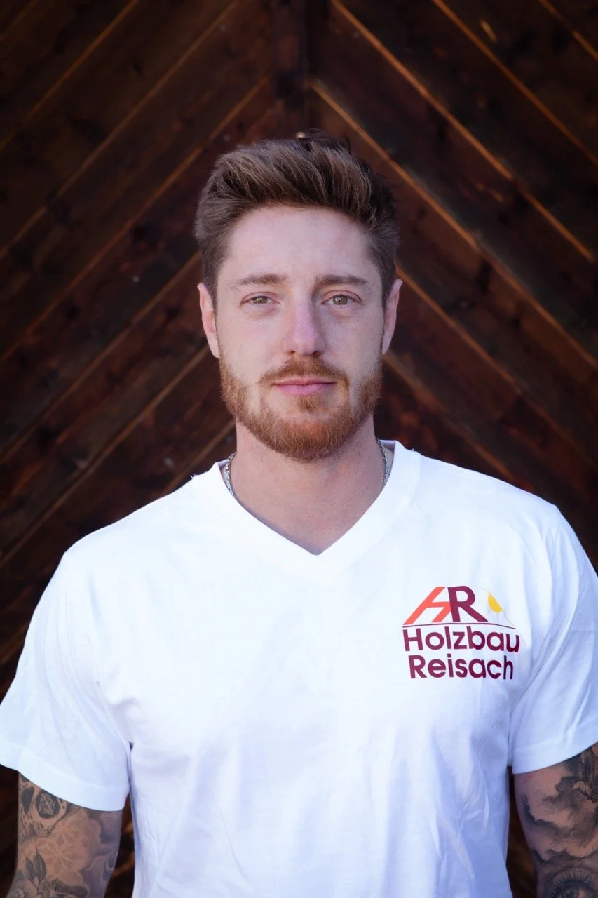 Junge Mann mit roten Haaren und Bart trägt weißes T-Shirt mit Logo und Text "Holzbau Reisch" vor Holzwand