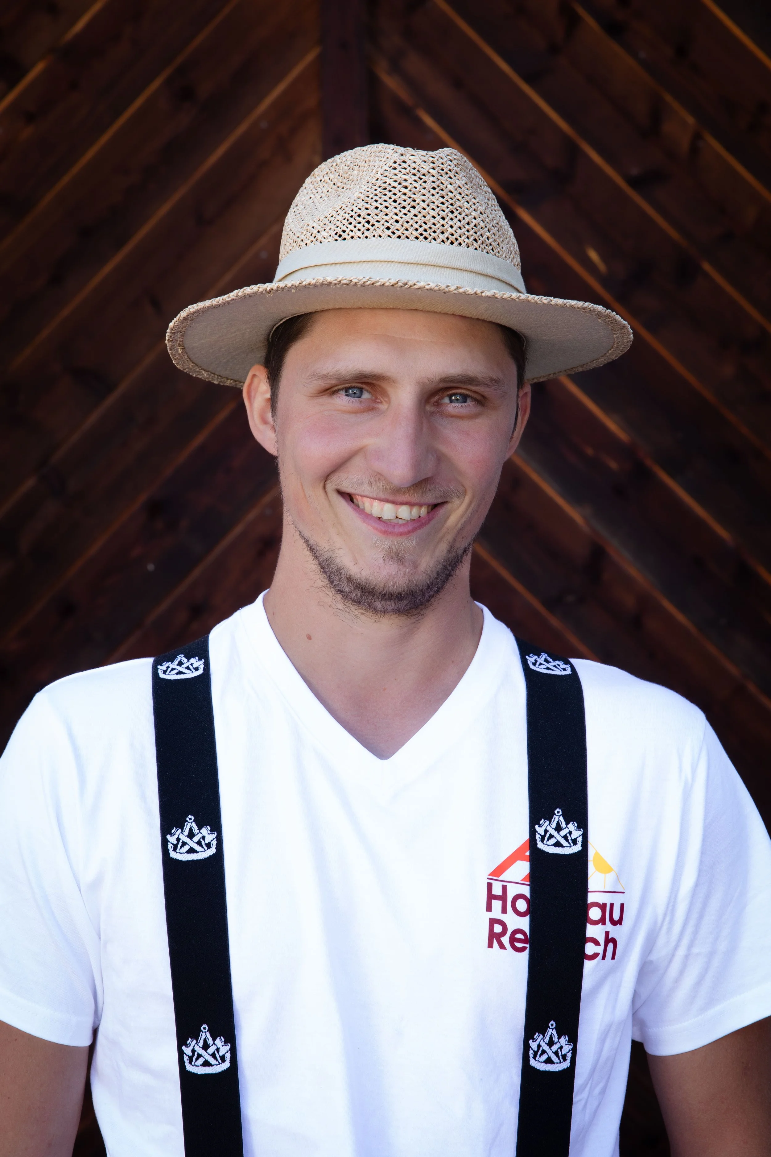 Lächelnder junger Mann mit blauen Augen, trägt einen Strohhut und ein weißes T-Shirt mit einem Logo und schwarzen Suspendern, im Hintergrund eine dunkle Holzdecke.