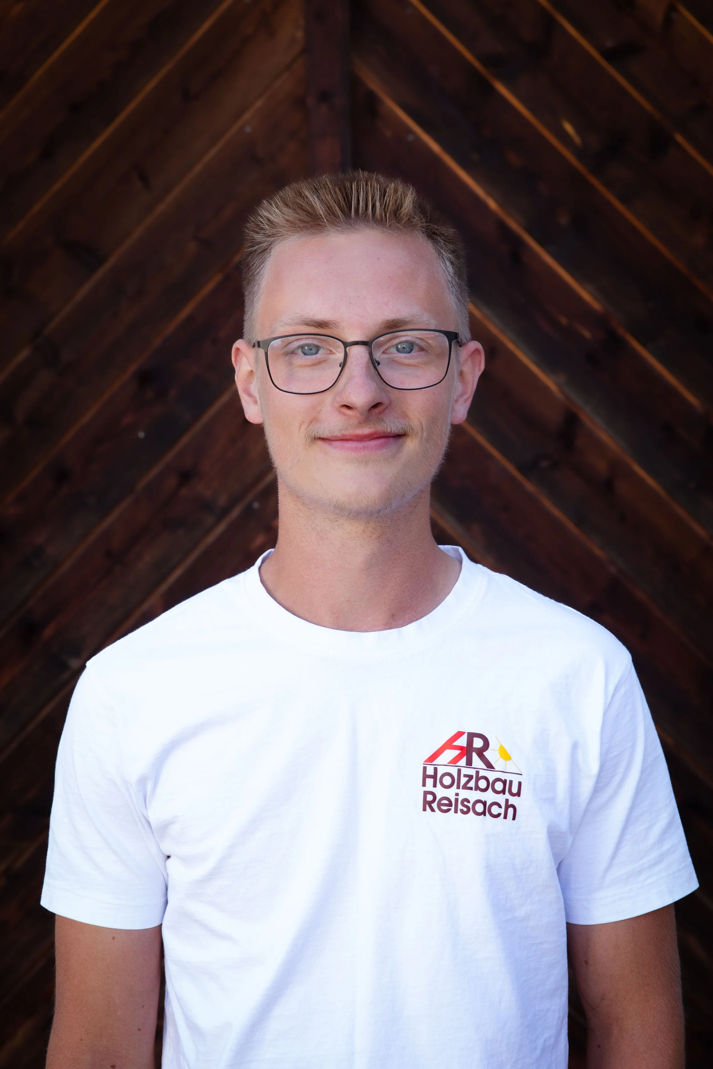 Junge Mann mit Brille trägt weißes T-Shirt mit Firmenlogo, stehend vor einer dunklen Holzdecke.