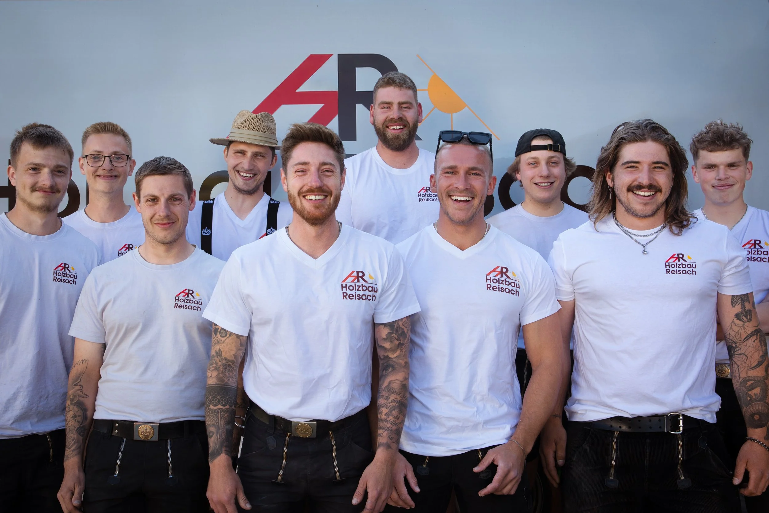 Ein Gruppenfoto von zwölf jungen Männern, die alle weiße T-Shirts mit dem Logo 'AR Holzbau Reischach' tragen. Sie stehen vor einer Wand mit einem großen Logo, das die Buchstaben 'AR' und einen Sonnen- und Holz-Design zeigt. Die Männer lachen und posieren für die Kamera.