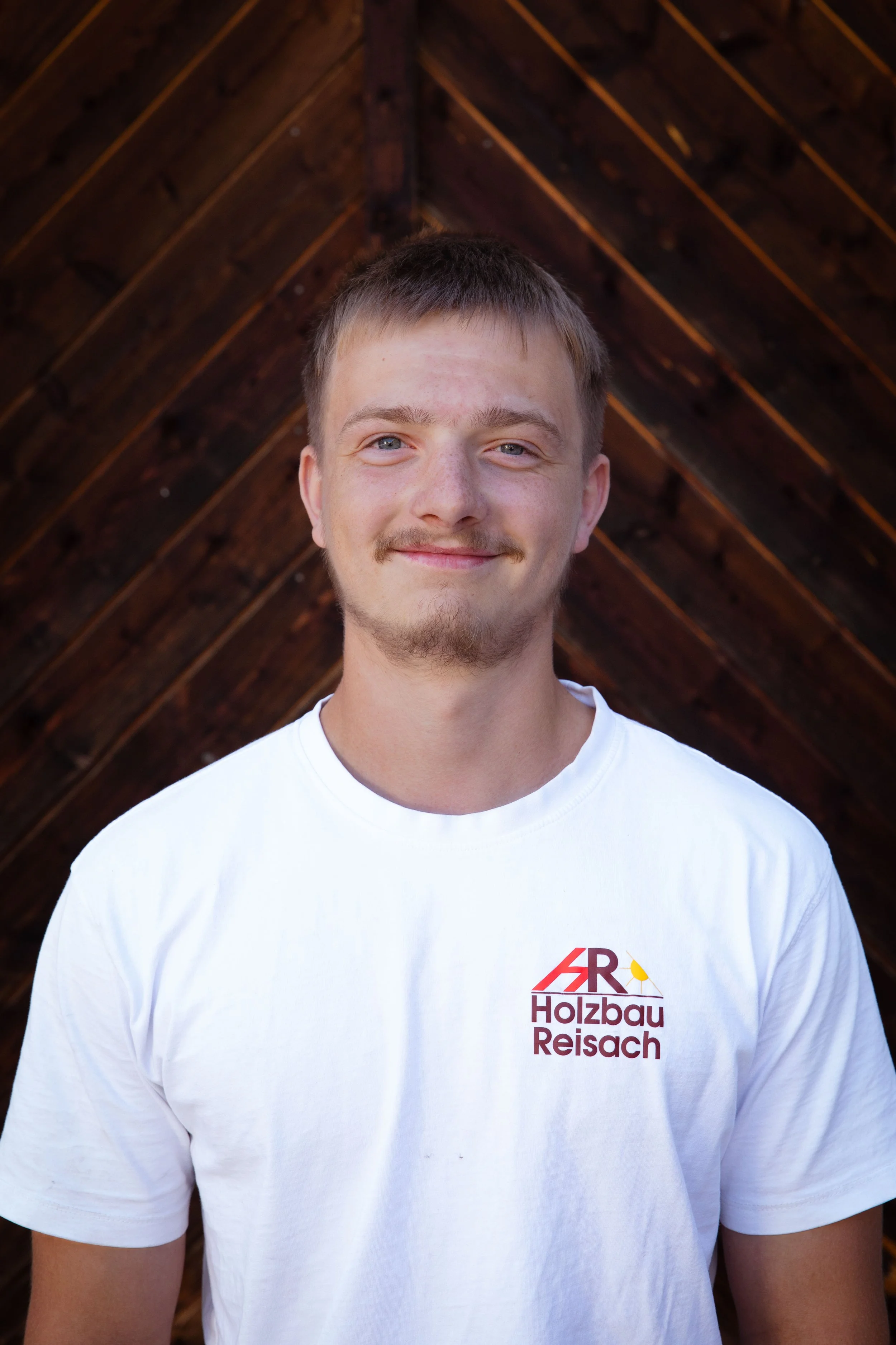 Junge trägt weißes T-Shirt mit Logo und Text 'Holzbau Reisch' vor einer Holzwand