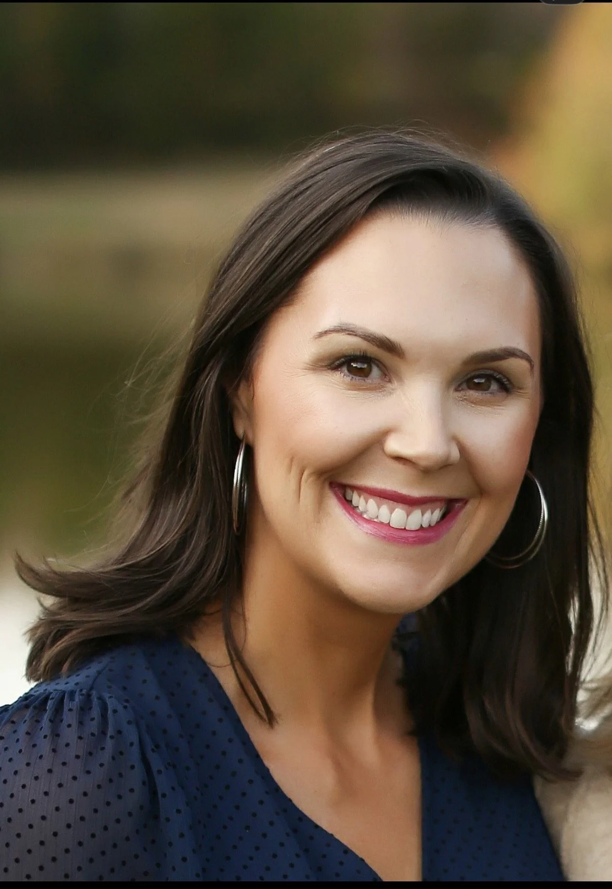 Close-up of a woman with dark brown hair, smiling, wearing earrings and a blue polka-dot top, outdoors with a blurred background.