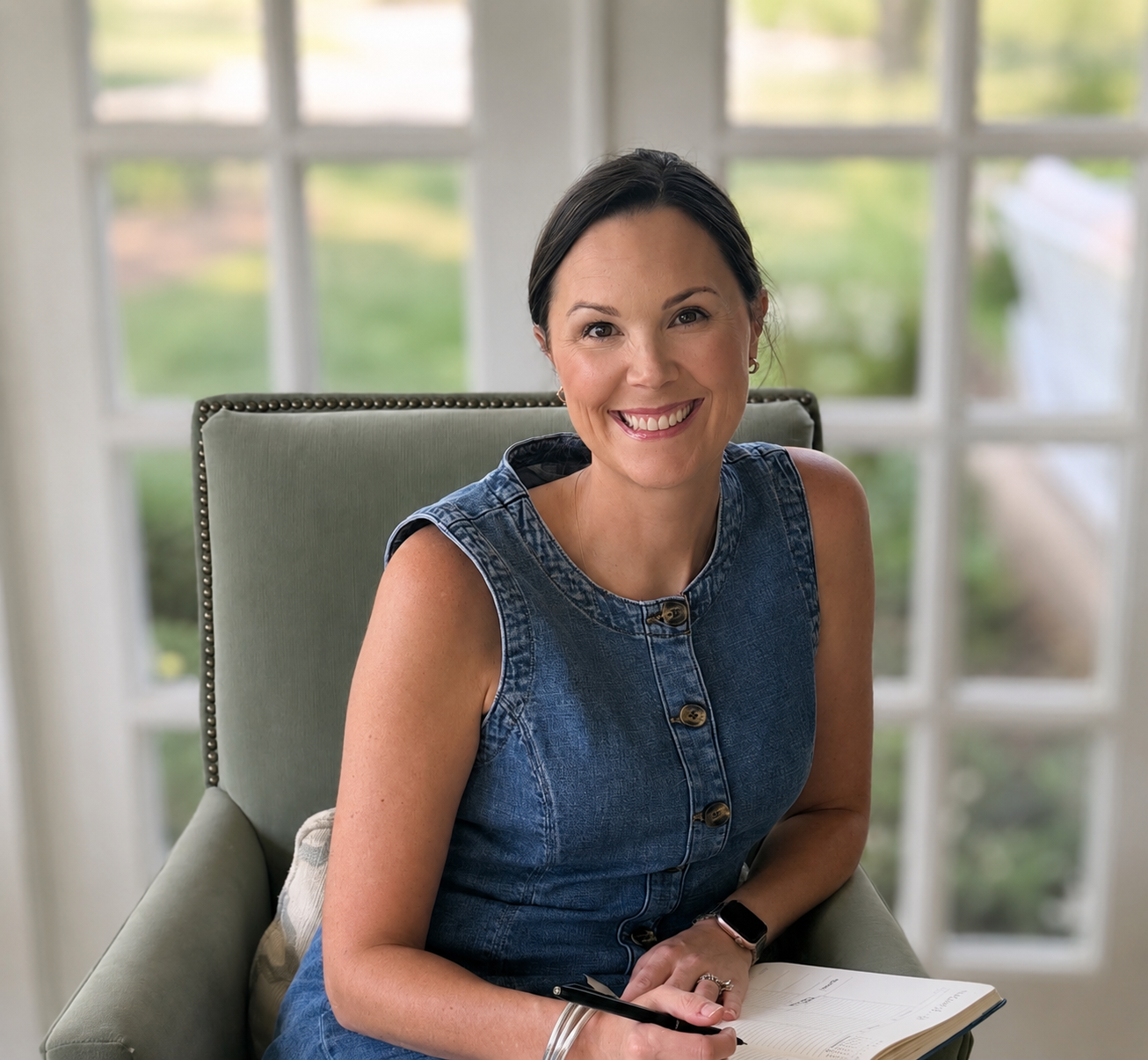 Woman smiling and sitting on a green chair near a window, holding a pen and a notebook.