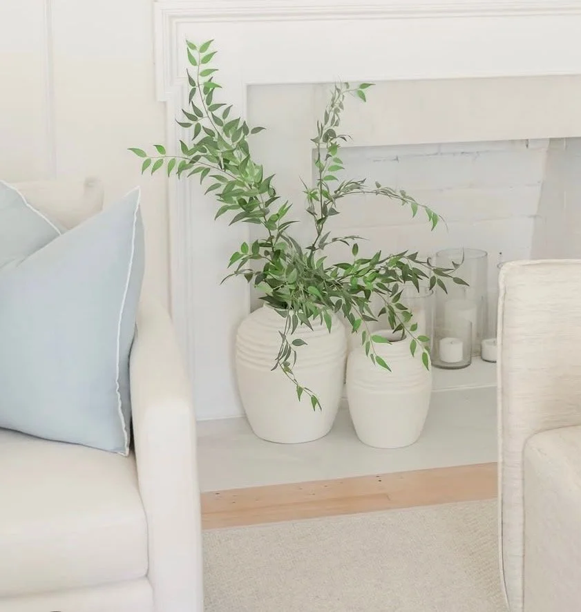 Close-up of a green leafy plant in a white ceramic pot placed on the floor between a white chair with a pale blue pillow and a beige upholstered piece of furniture, with additional candles and glass containers in the background.