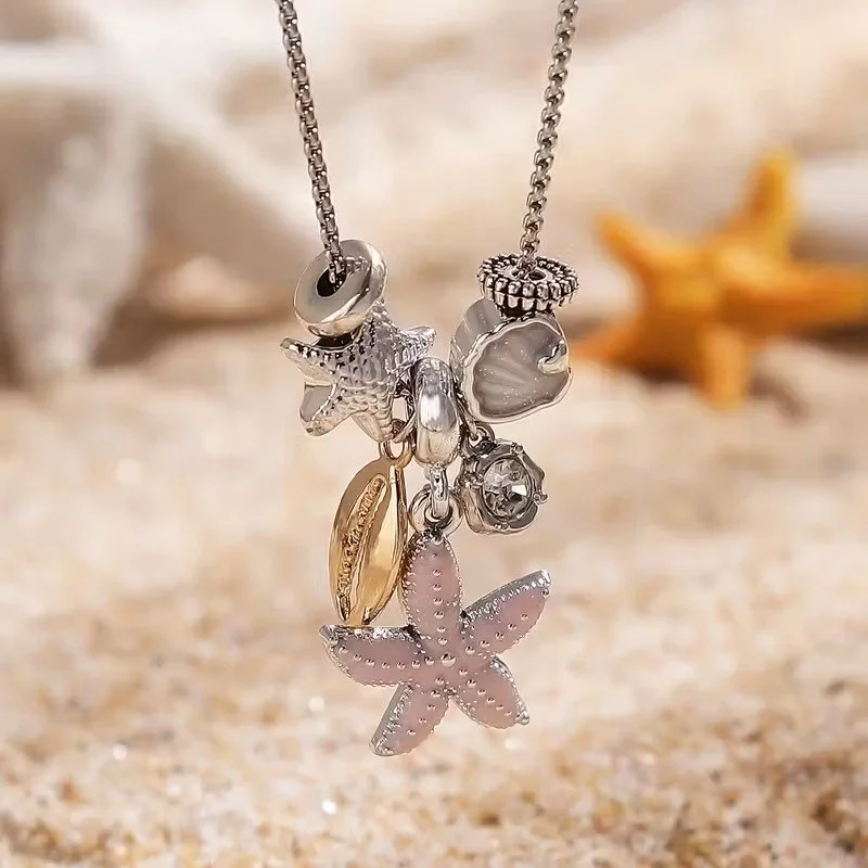Silver necklace with multiple pendant charms, including a pink flower, gold leaf, starfish, shell, bracelet, and small gemstone, placed on sandy surface with starfish in background.