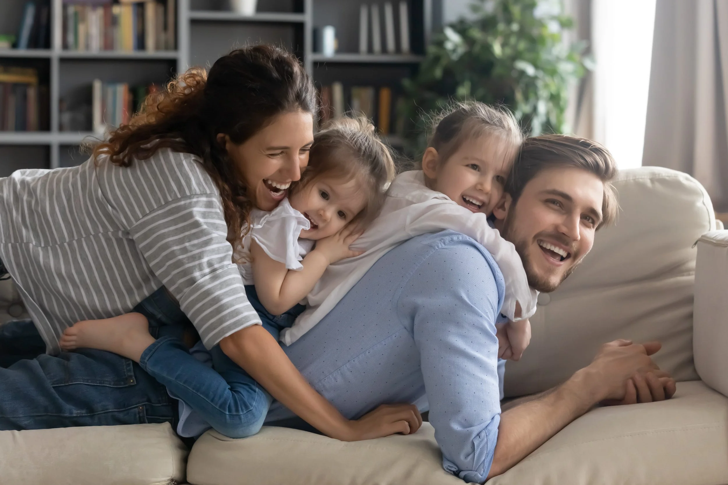 A family of fou, including a mother, father, and two young girls, playing and laughing together on a sofa in a cozy living room.