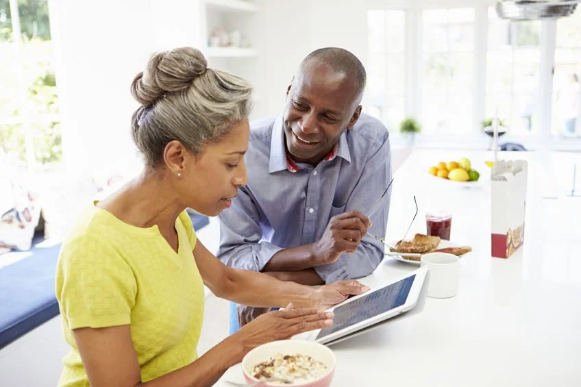 An older woman and a man are having breakfast in a bright kitchen, looking at a tablet together. The woman is in a yellow shirt, and the man is in a blue shirt. There are food and drinks on the kitchen counter.