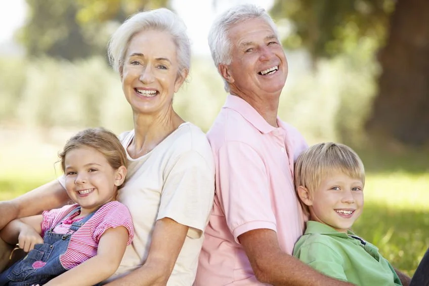 Senior grandparents and grandchildren sitting outdoors on grass, smiling and enjoying the sunny day.
