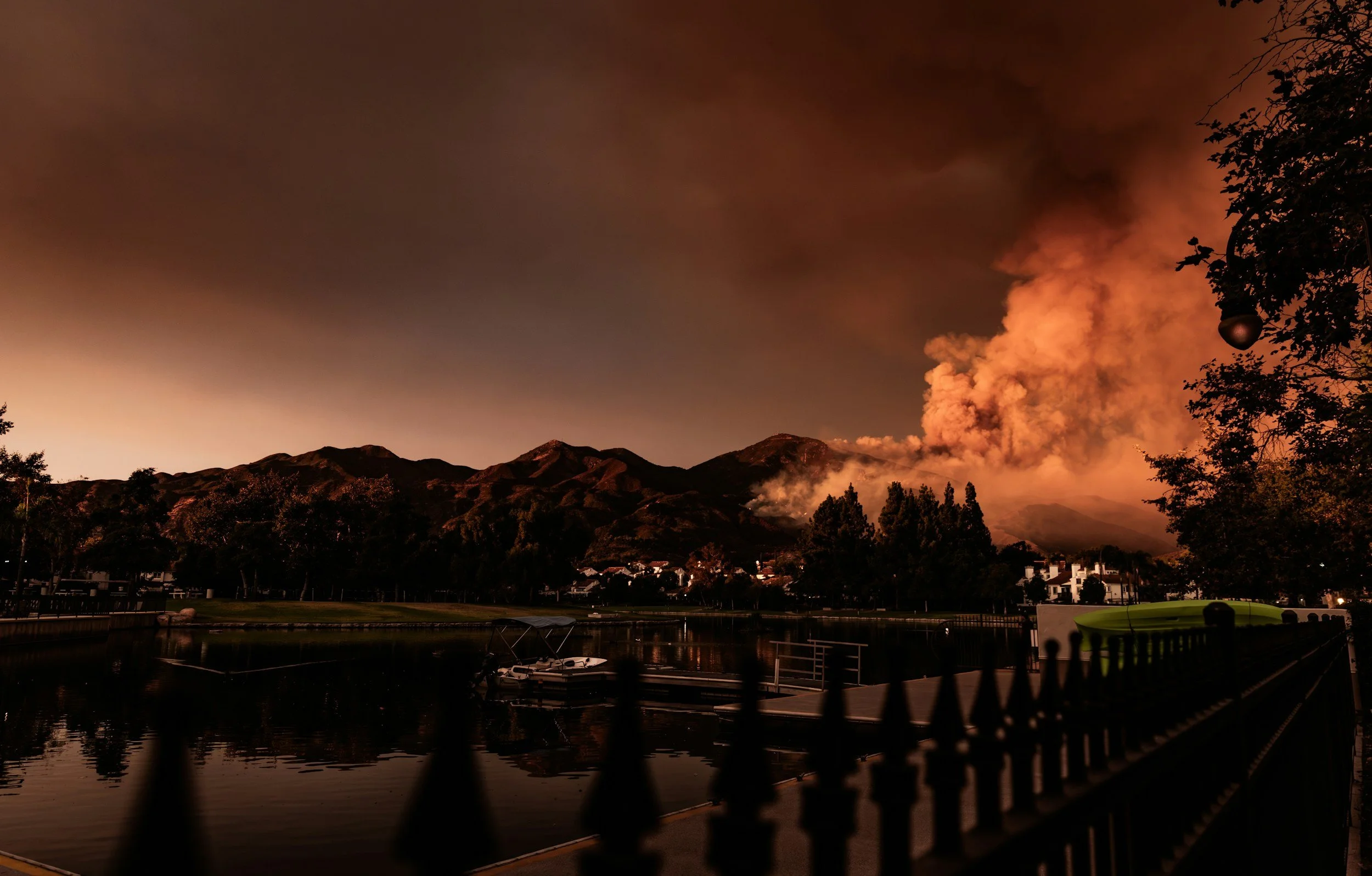 Hills and mountains with smoke and clouds in the sky at dusk, near a body of water with boats and houses, and trees on the right.