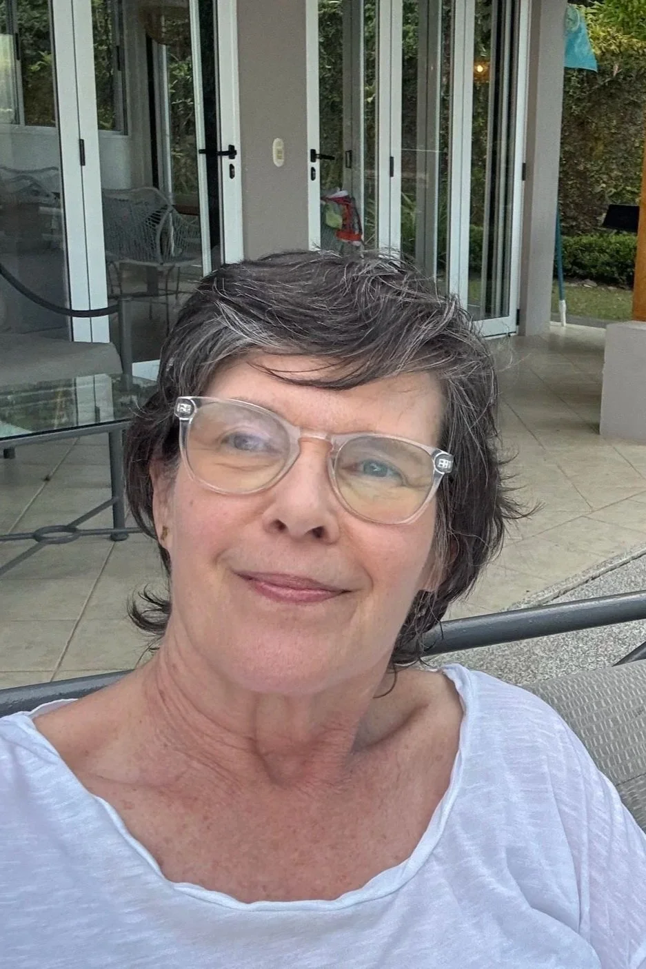 A smiling woman with short gray hair and glasses, sitting outdoors on a patio with a glass table and chairs, behind her a house with open sliding glass doors.
