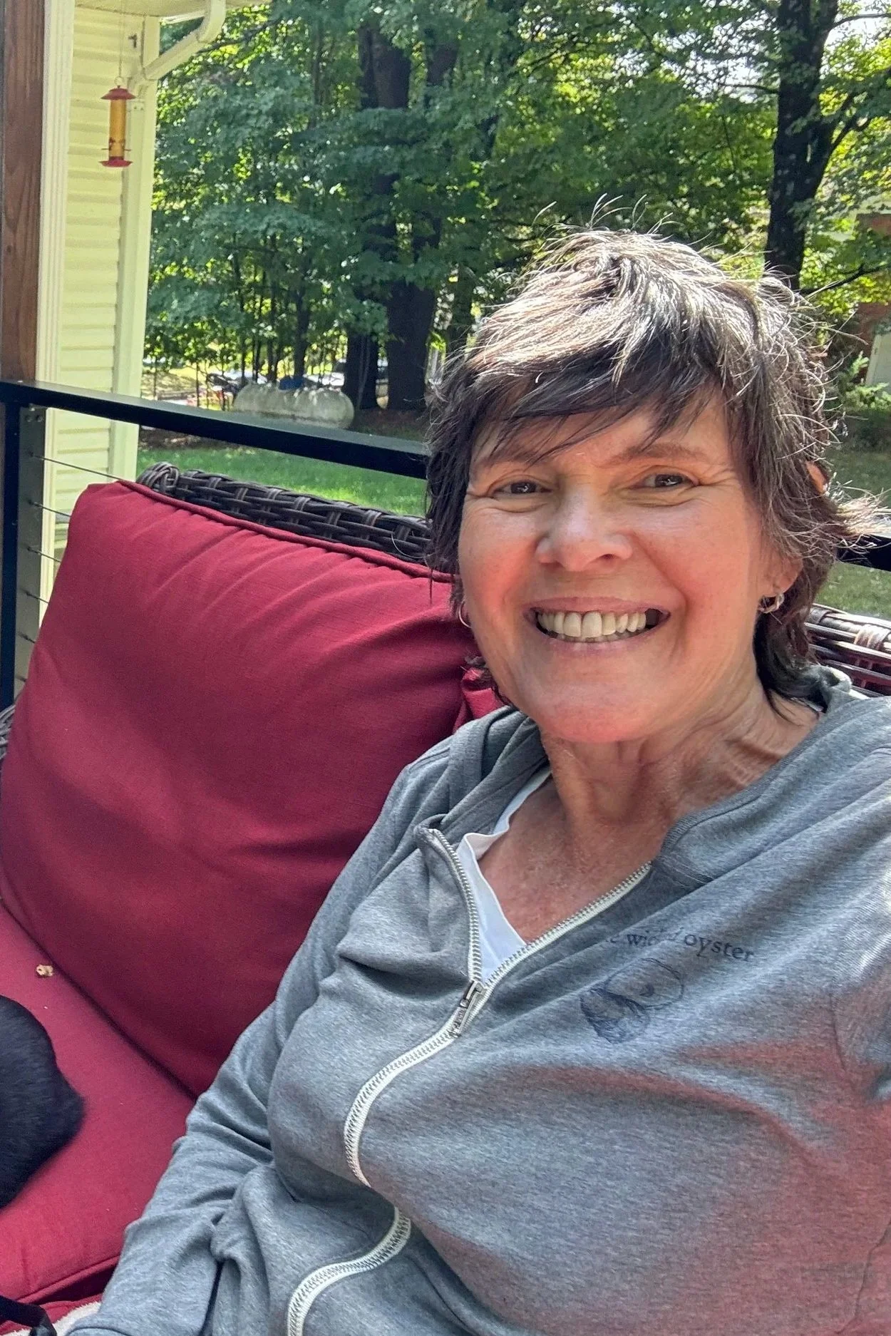 Smiling woman with short gray hair sitting on a red outdoor cushion on a patio with trees in the background.