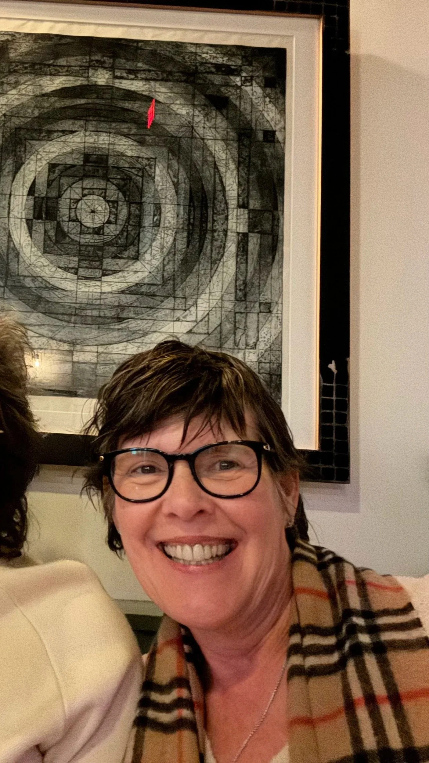 A smiling woman with glasses and short hair taking a selfie with a framed geometric art piece in the background.