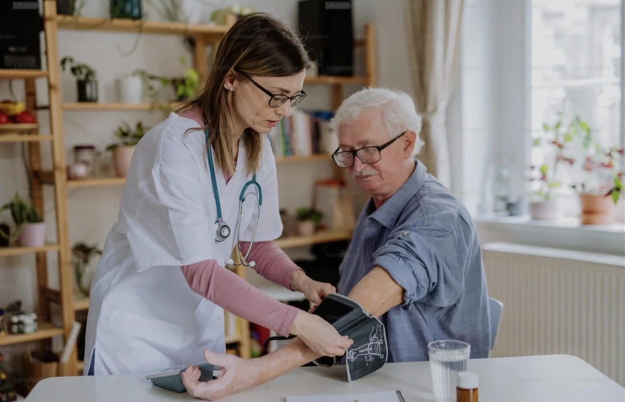 A female healthcare professional in a white coat checks the blood pressure of an elderly man in a bright room with shelves and plants.