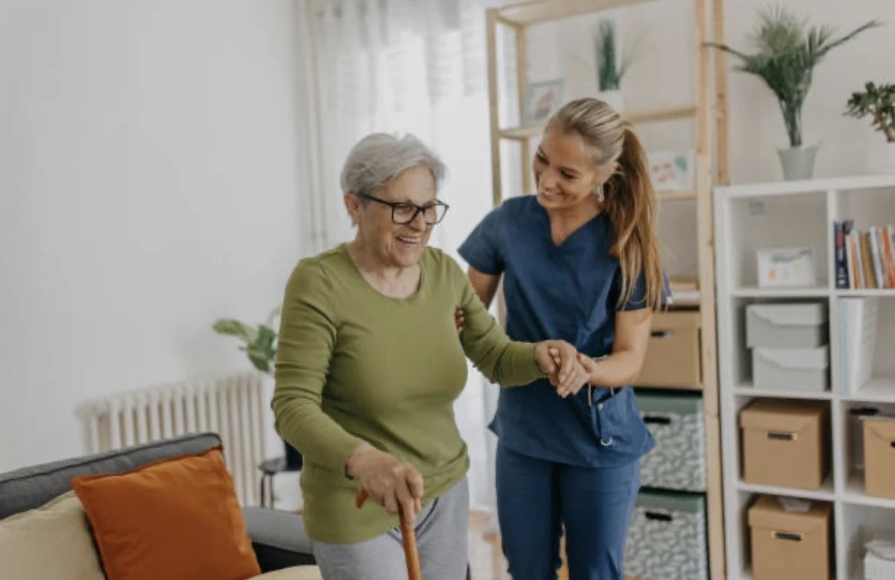 A young female caregiver assisting an elderly woman with walking in a living room, both smiling and holding hands.