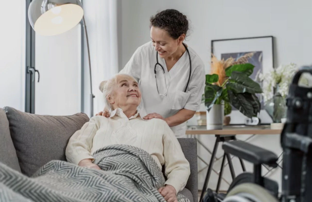 A healthcare worker, wearing white, is smiling and leaning over a senior woman sitting on a couch, who is smiling up at her. The elderly woman is covered with a patterned blanket and appears to be receiving care or a visit at home, with a camera visible in the foreground.
