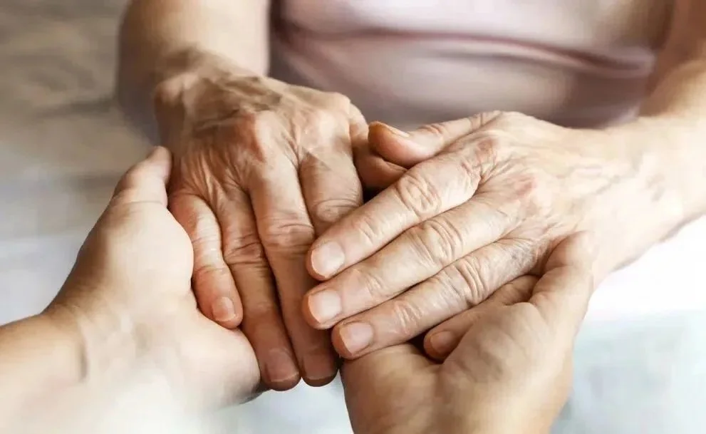 Two pairs of hands, one adult and one elderly, holding each other in a gentle, supportive manner.