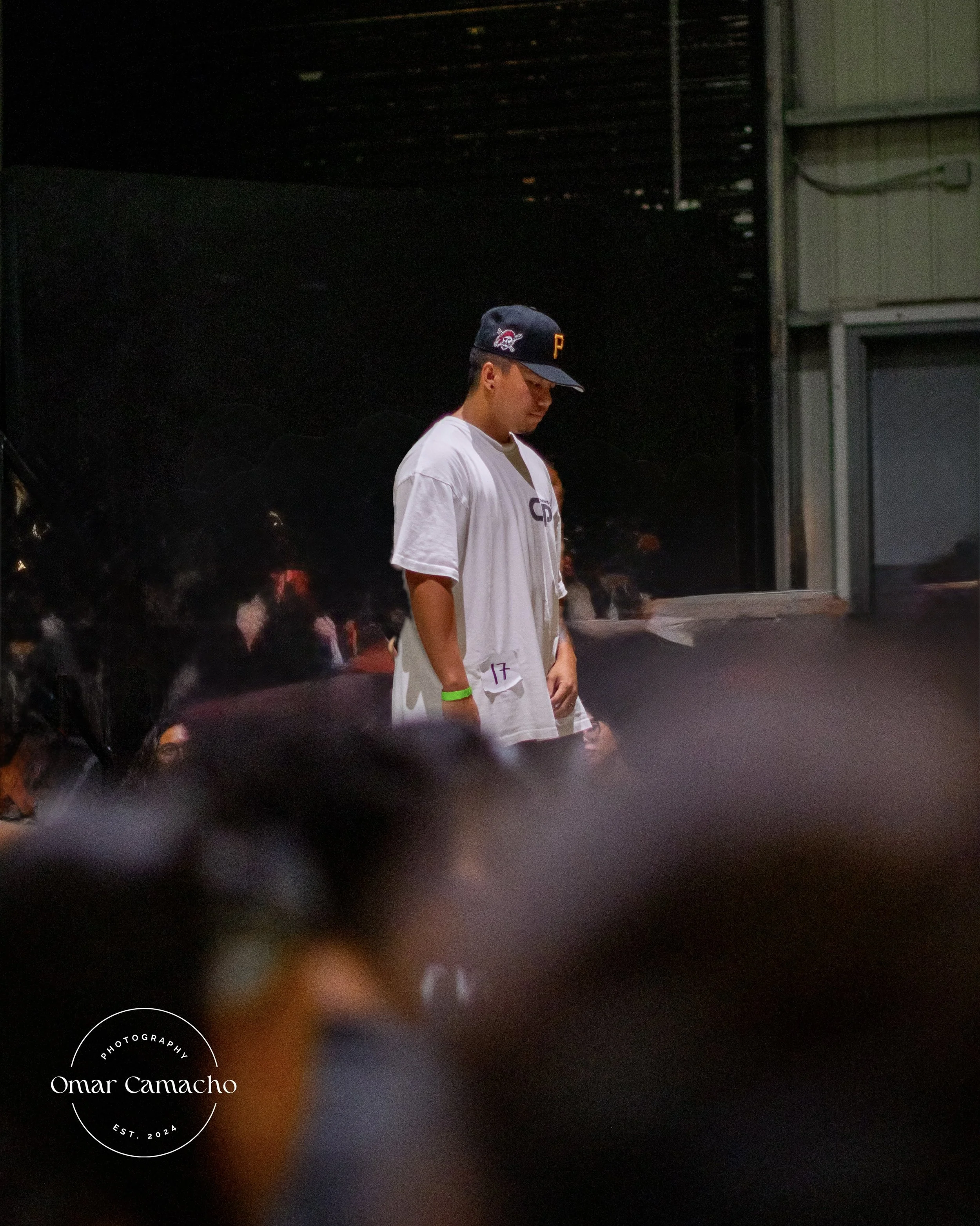 A young man wearing a cap and a white sports jersey stands with head bowed in a dimly lit room with a dark wall and some people behind him.