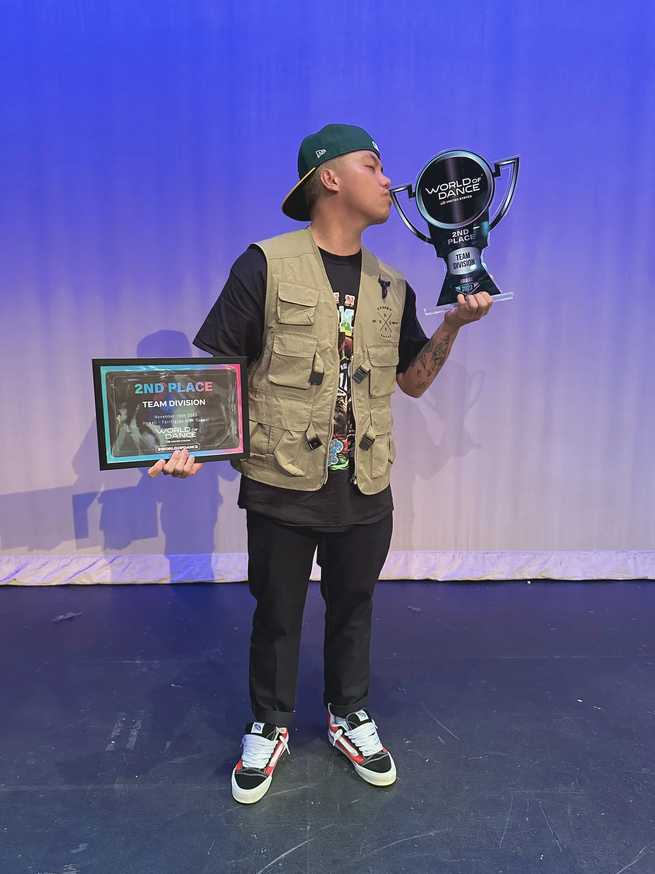 Man wearing a green hat and beige vest holding a trophy and an award plaque, standing on a stage with purple lighting.