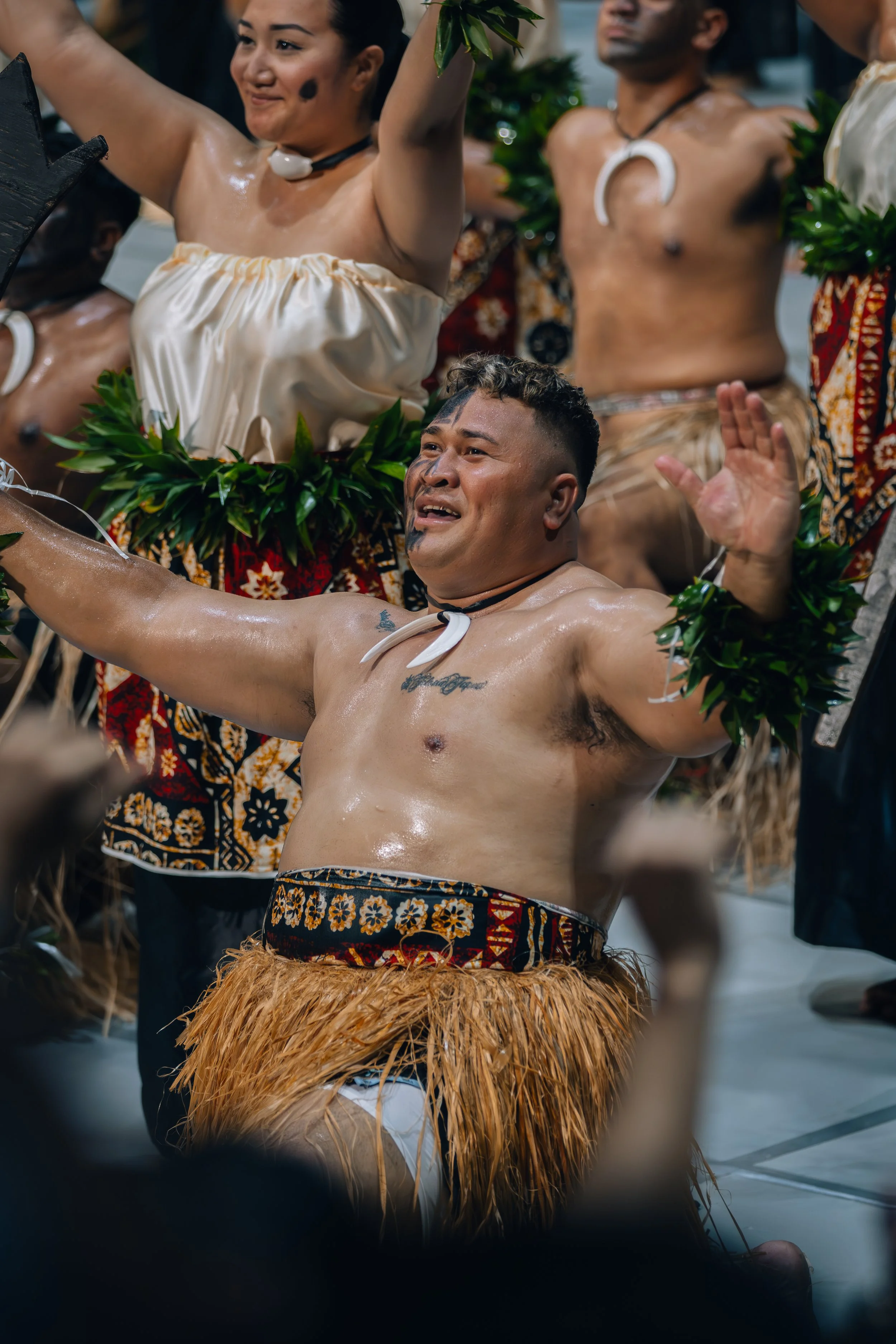 A group of men and women in traditional Polynesian attire participating in a cultural dance or ceremony. The central figure, a man, is kneeling with arms raised, wearing a grass skirt, with a joyful expression. Others are standing behind him, some wi