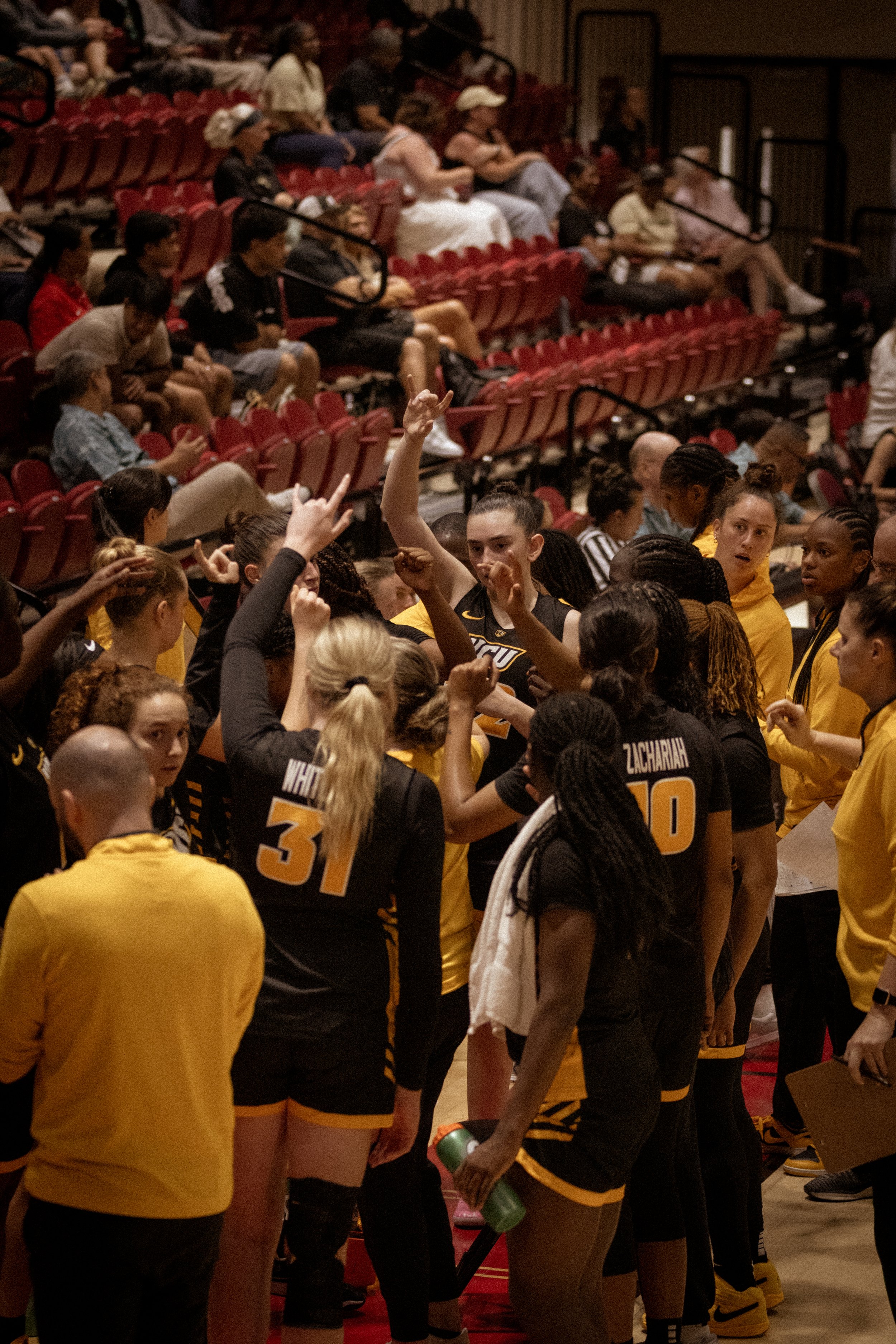 A group of female basketball players in black and yellow uniforms gather together on a basketball court, some with fists raised, and are surrounded by a few staff members, while spectators sit in red seats in the background.