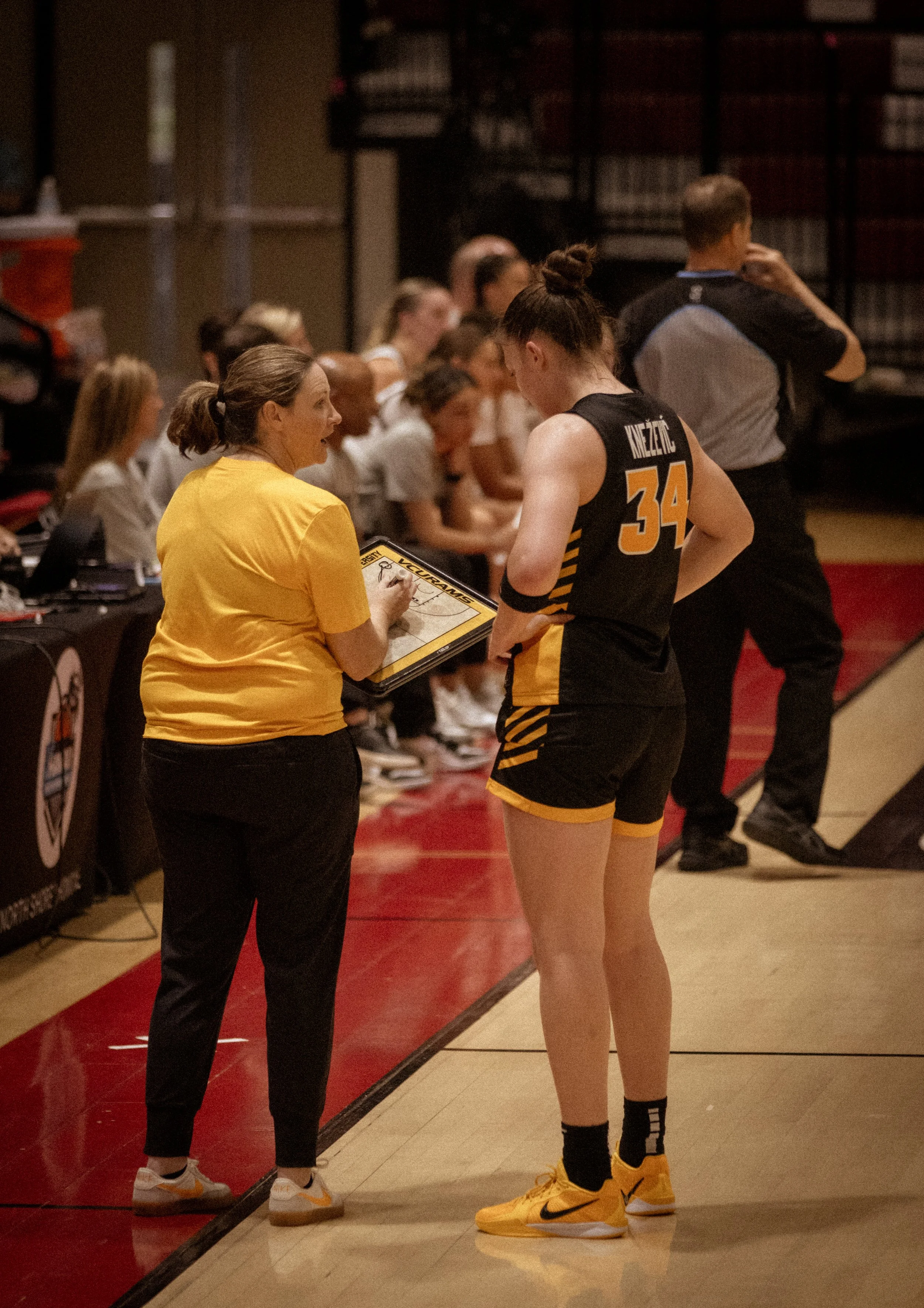 A female basketball player in a black and yellow uniform standing on a basketball court during a game, speaking with a female coach who is holding a clipboard. Other players are seated on the bench in the background, with a referee in a gray and blac