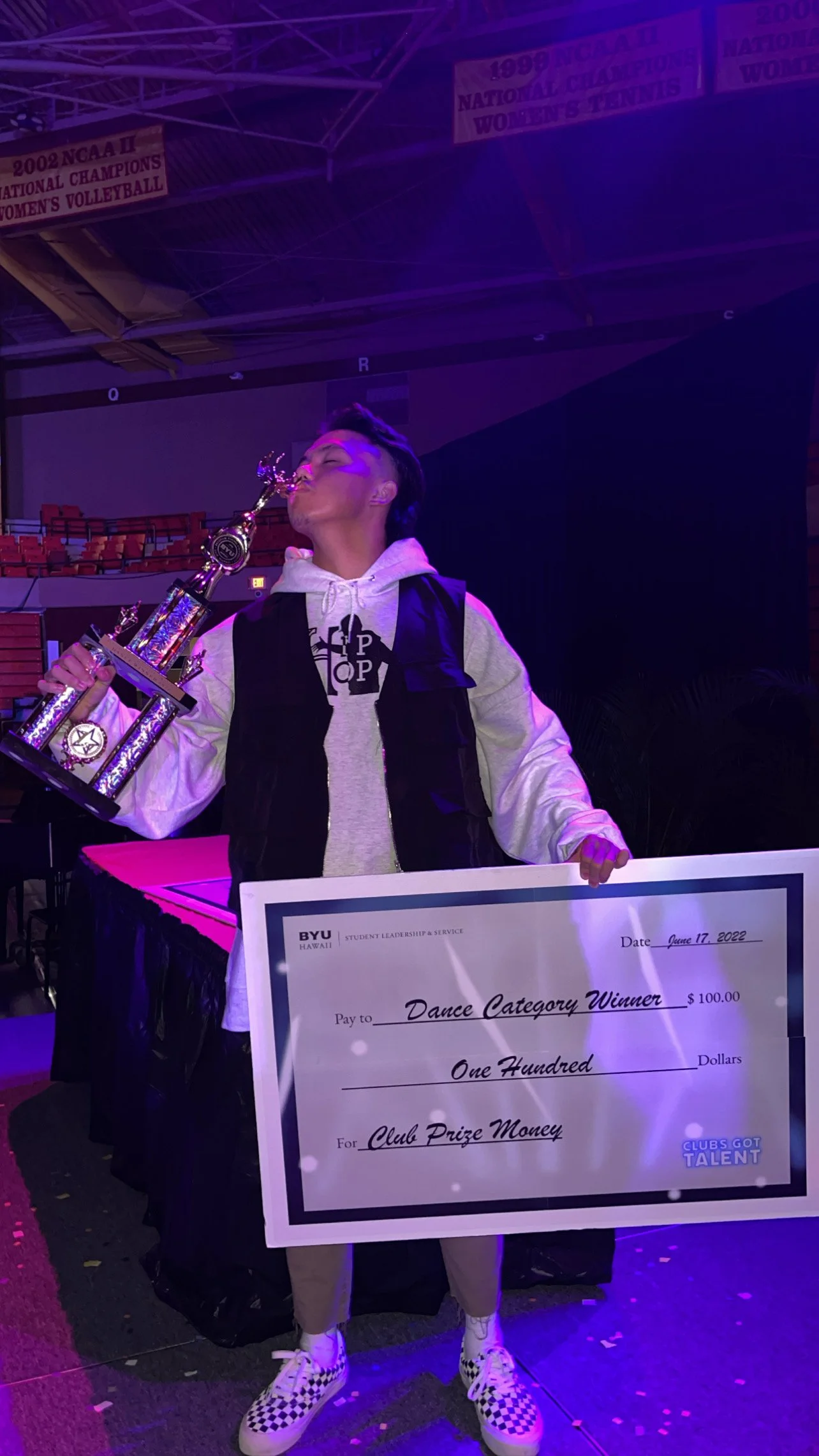 A person holding a trophy and a giant check for $100, awarded as the dance category winner at a Boise State University event on June 17, 2022. The person is standing in an indoor arena, wearing checkered shoes and a gray hoodie.