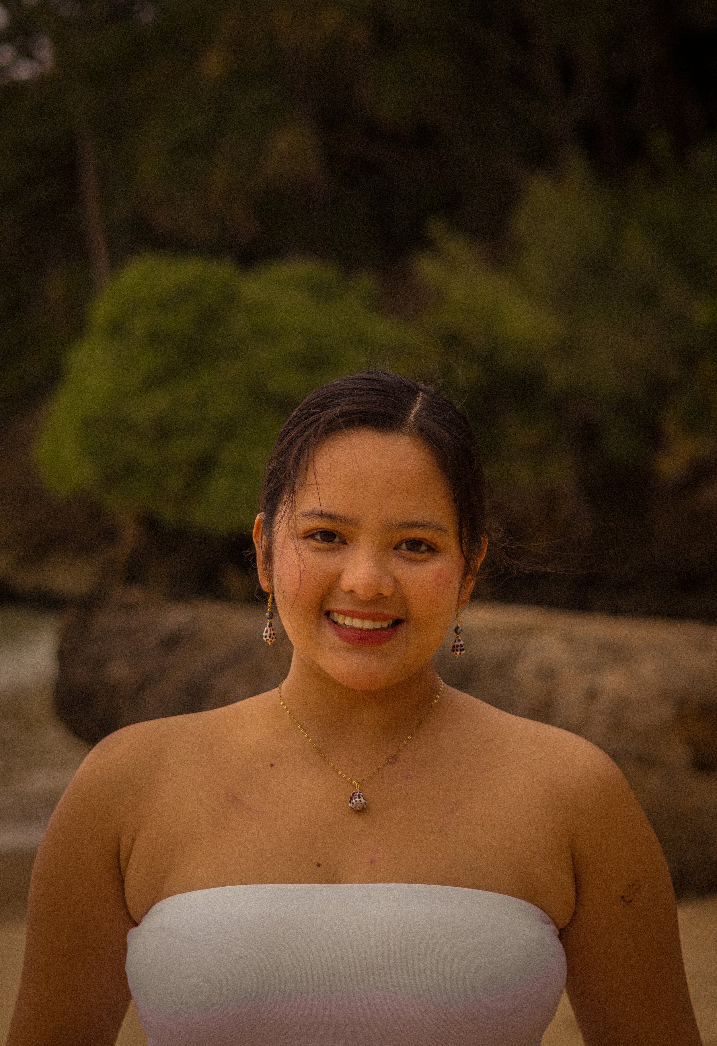 A smiling young woman with dark hair tied back, wearing a white strapless top and jewelry, standing outdoors near rocks and greenery.