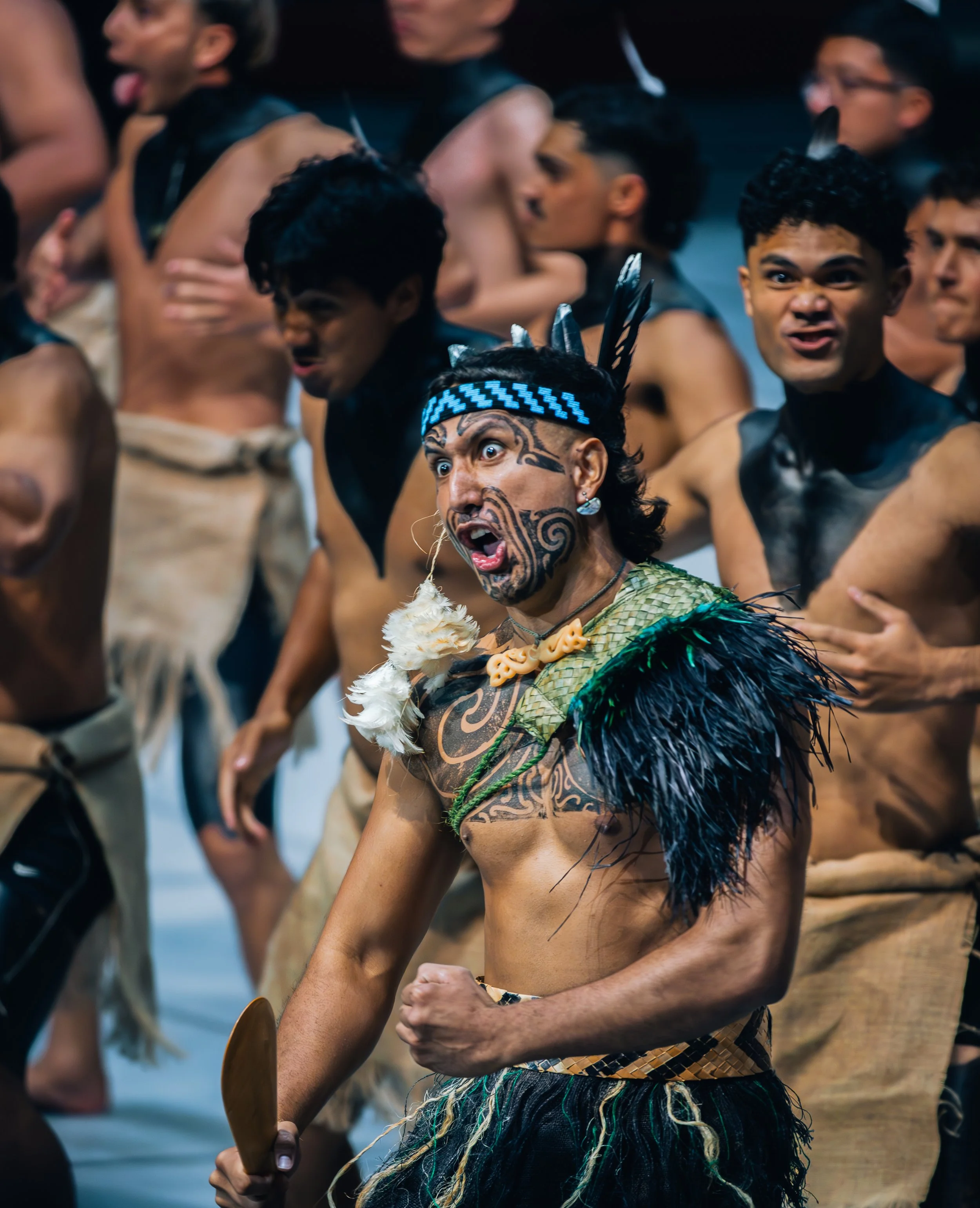 A man in traditional Polynesian attire, including a feathered headband, body paint, and a woven skirt, appears to be performing or leading a cultural dance with a group of men dressed similarly in the background.