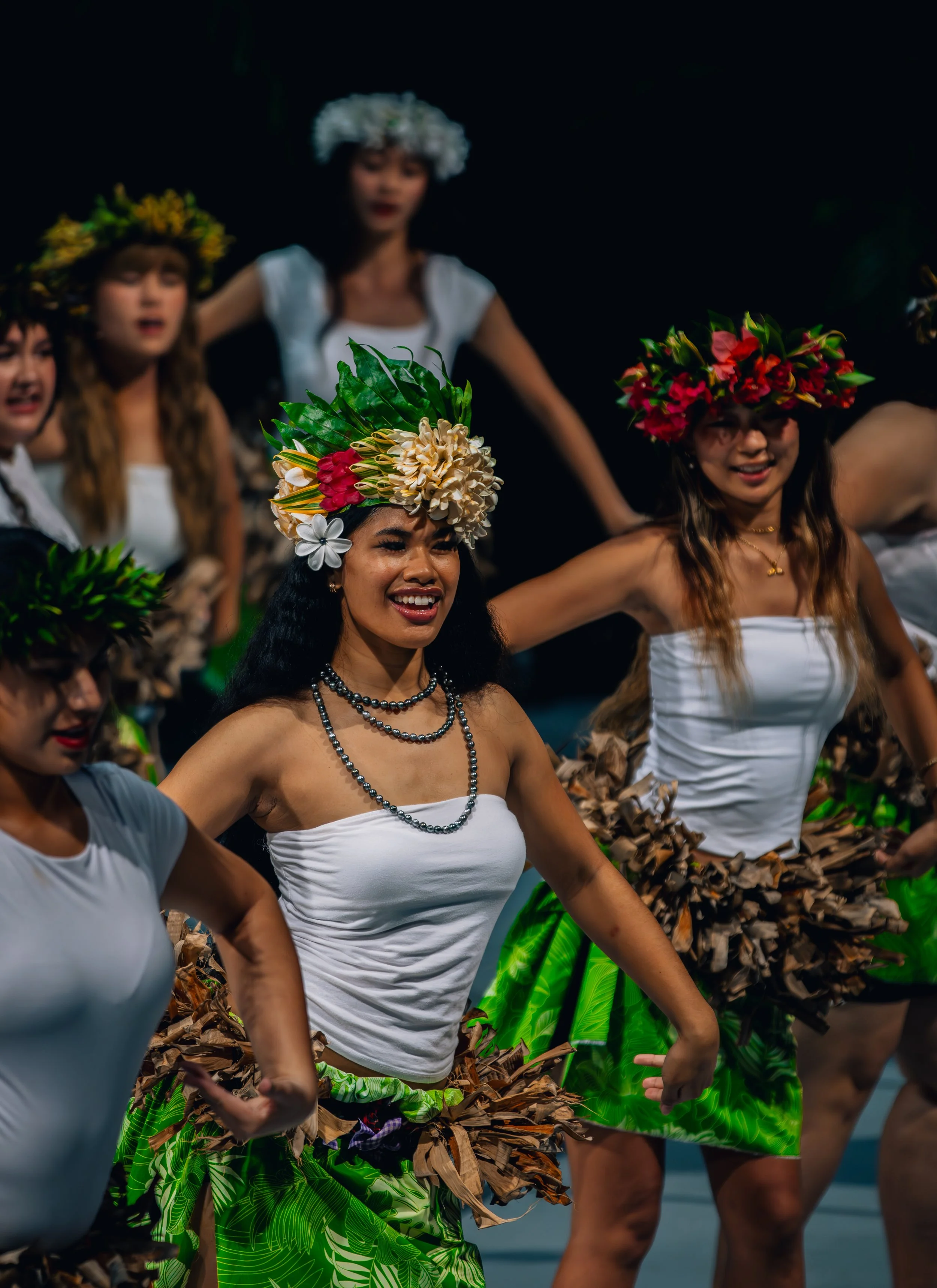 A group of women wearing traditional Hawaiian hula costumes with floral headpieces, grass skirts, and white tops, performing a dance on stage.