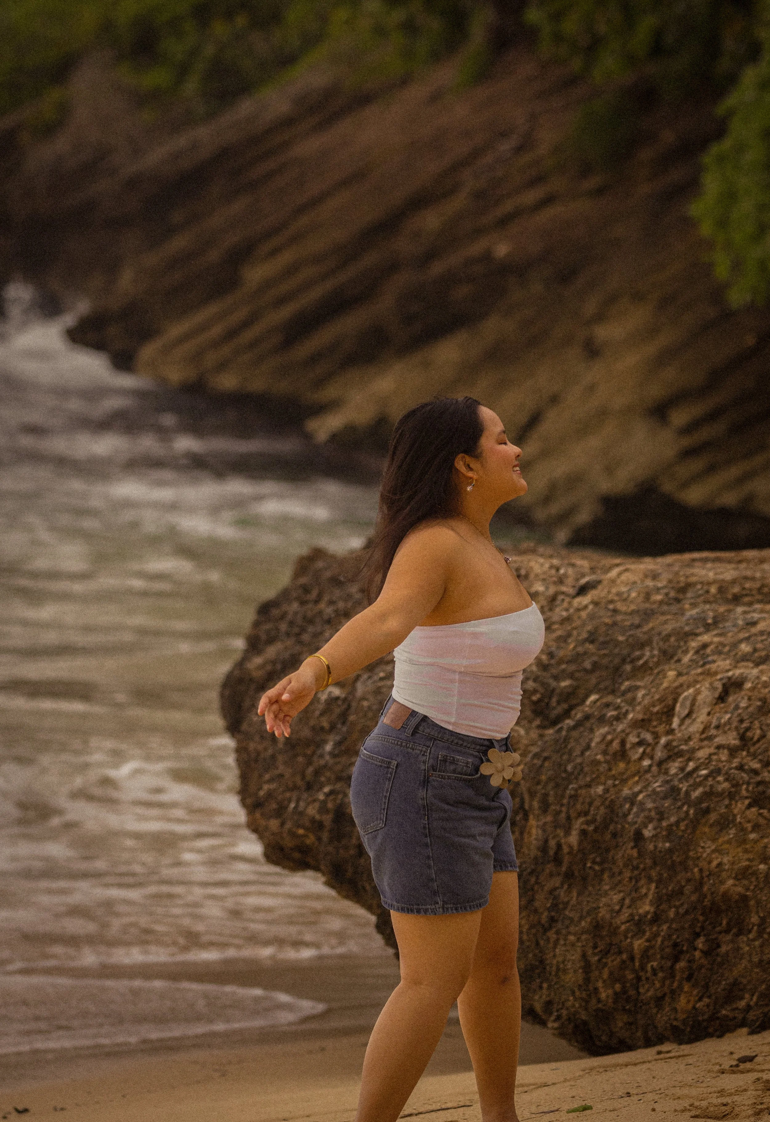 Woman standing on a beach with arms outstretched, smiling, near large rocks and a cliff at sunset.