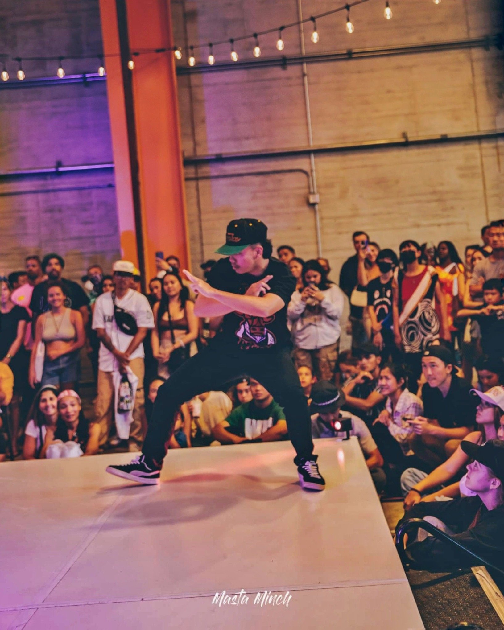 A person dancing on stage during a dance event with a crowd watching in an industrial-style venue with string lights hanging overhead.