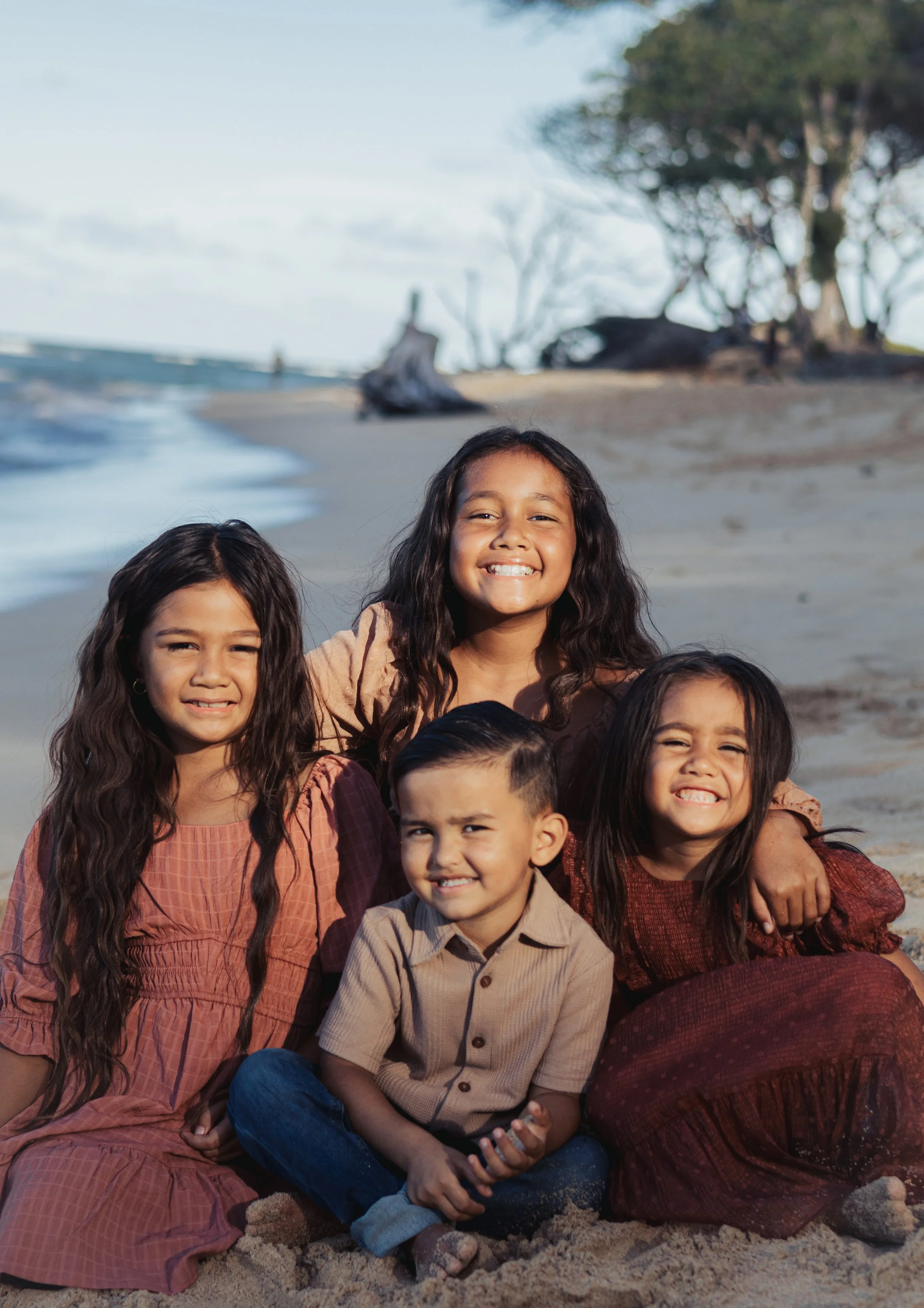Four children, three girls and one boy, sitting on the sand at the beach, smiling and enjoying the sunny day.