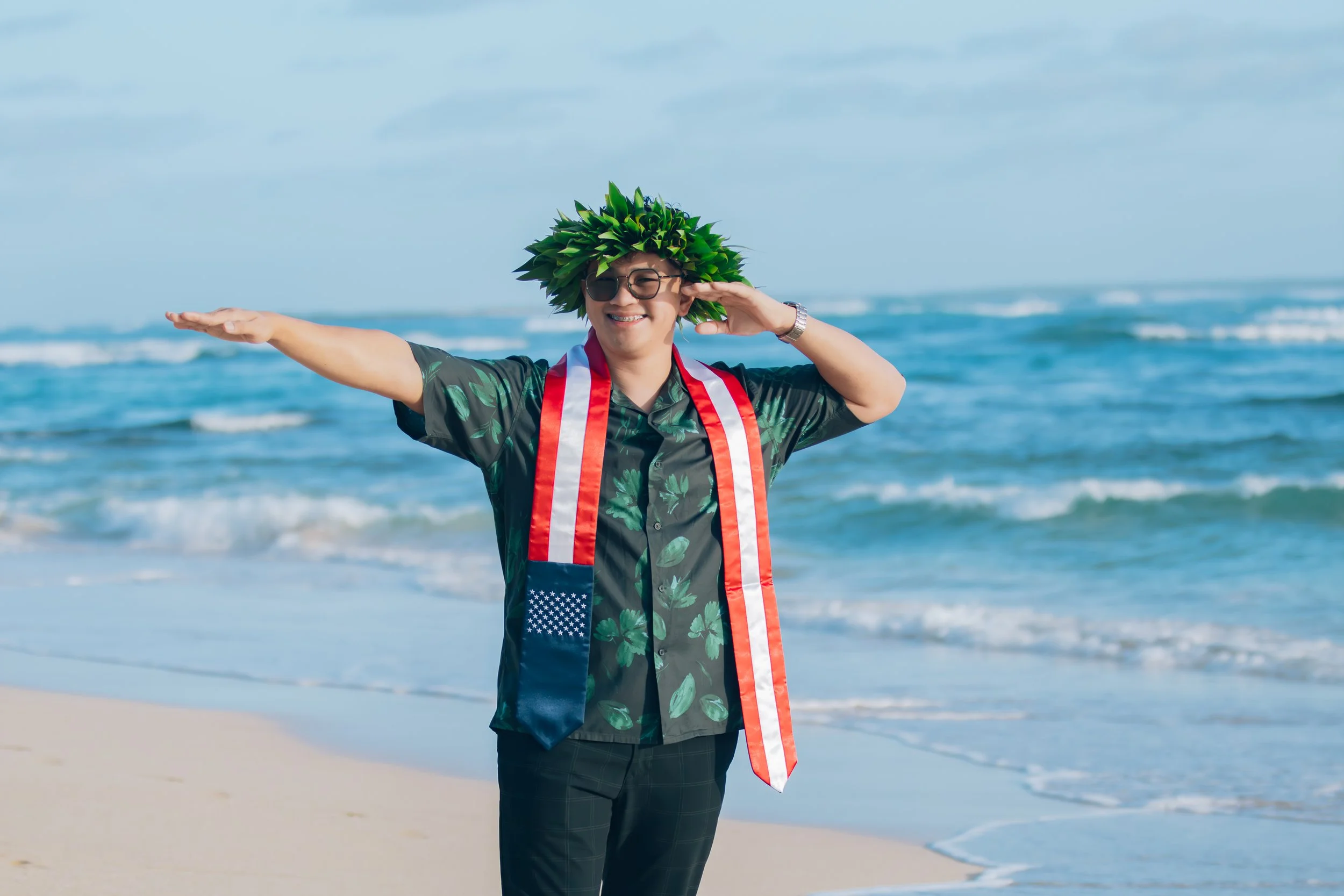 Smiling man on the beach wearing a leafy crown, sunglasses, a Hawaiian shirt, and a patriotic scarf, standing near the ocean with outstretched arms.