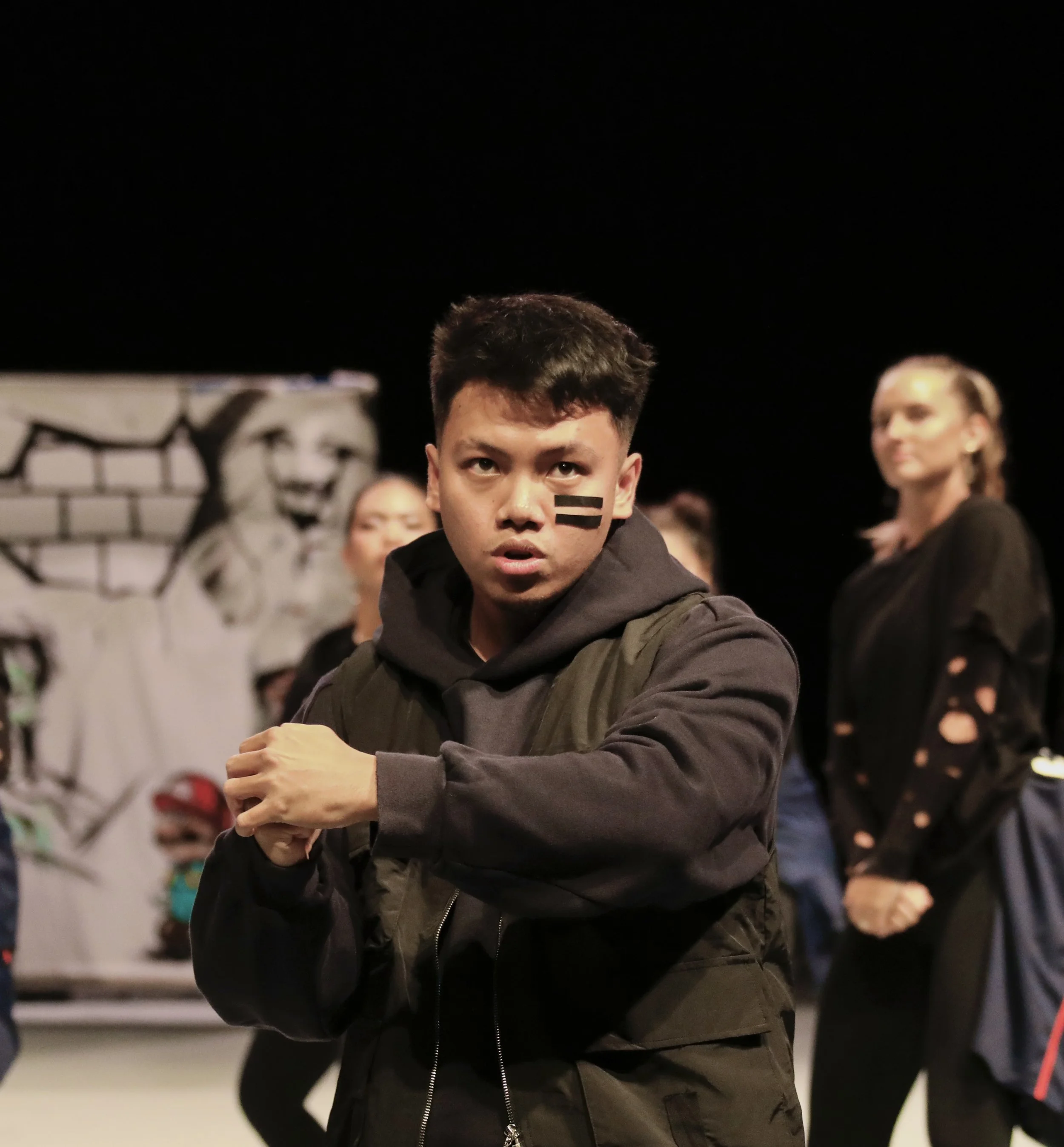 A young man with black face paint in a gym or performance space, performing a pose with clenched fists, with other people in the background and a mural of a woman.